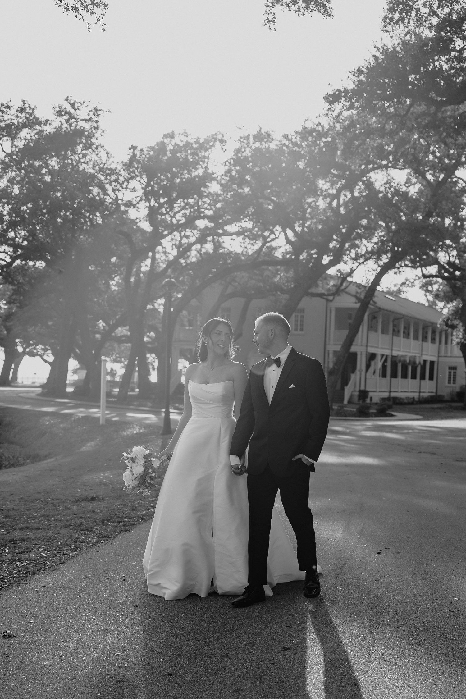 Black and white photo of a bride and groom holding hands on a tree-lined path on their wedding day.