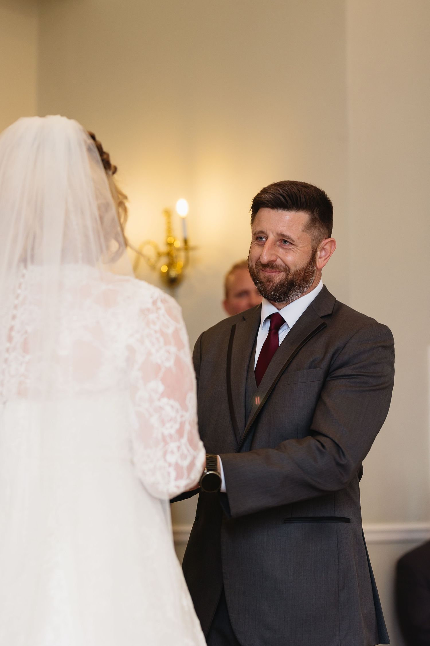 Groom with beard smiling warmly at bride in lace gown and veil at wedding altar.