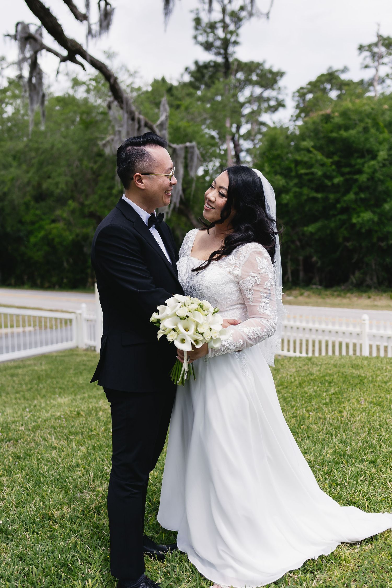 Asian bride and groom share romantic moment in lush garden, bride holding white calla lily bouquet in elegant lace wedding gown.