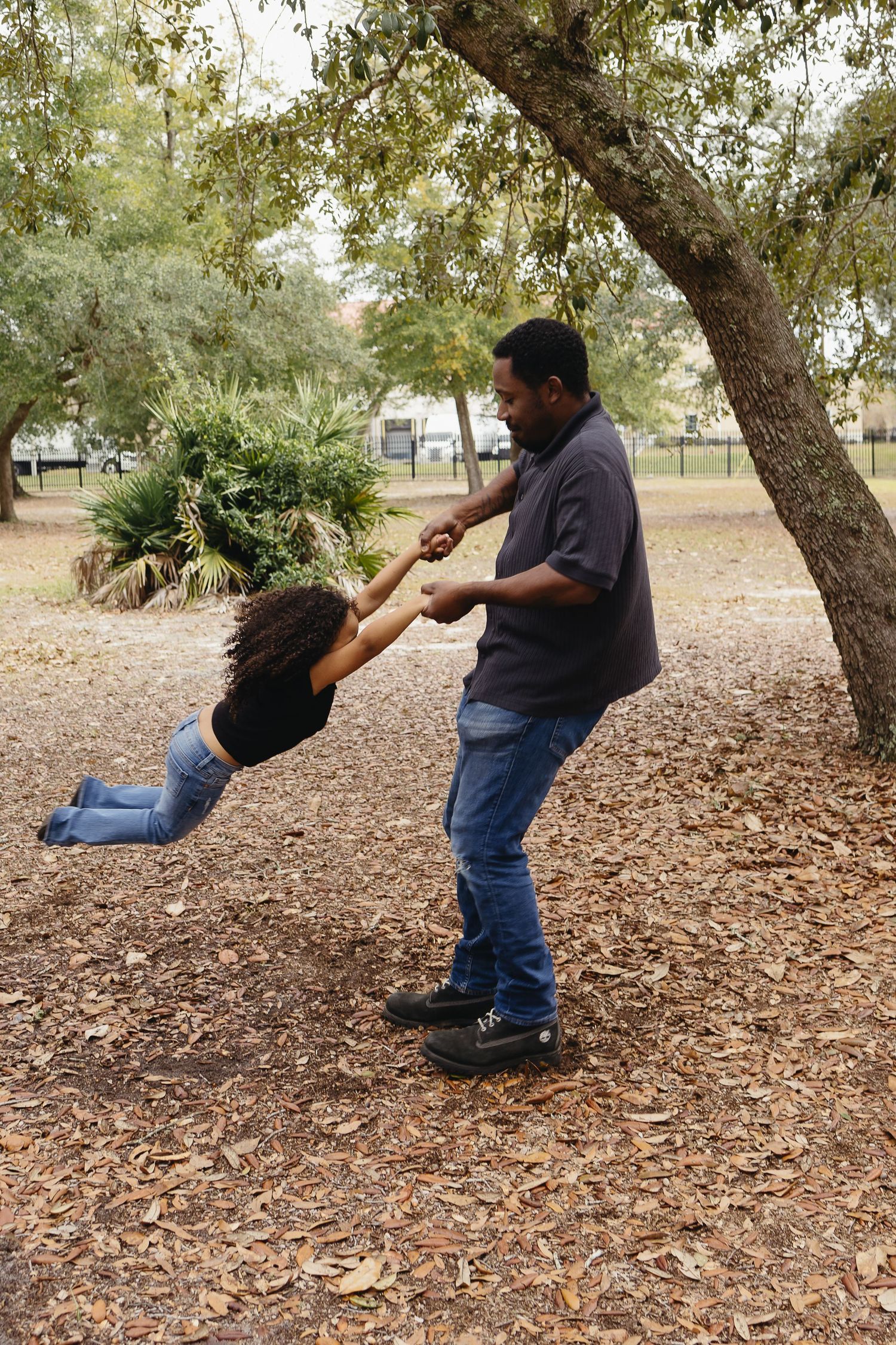A father playfully swinging his daughter by her hands under a tree in an outdoor park.