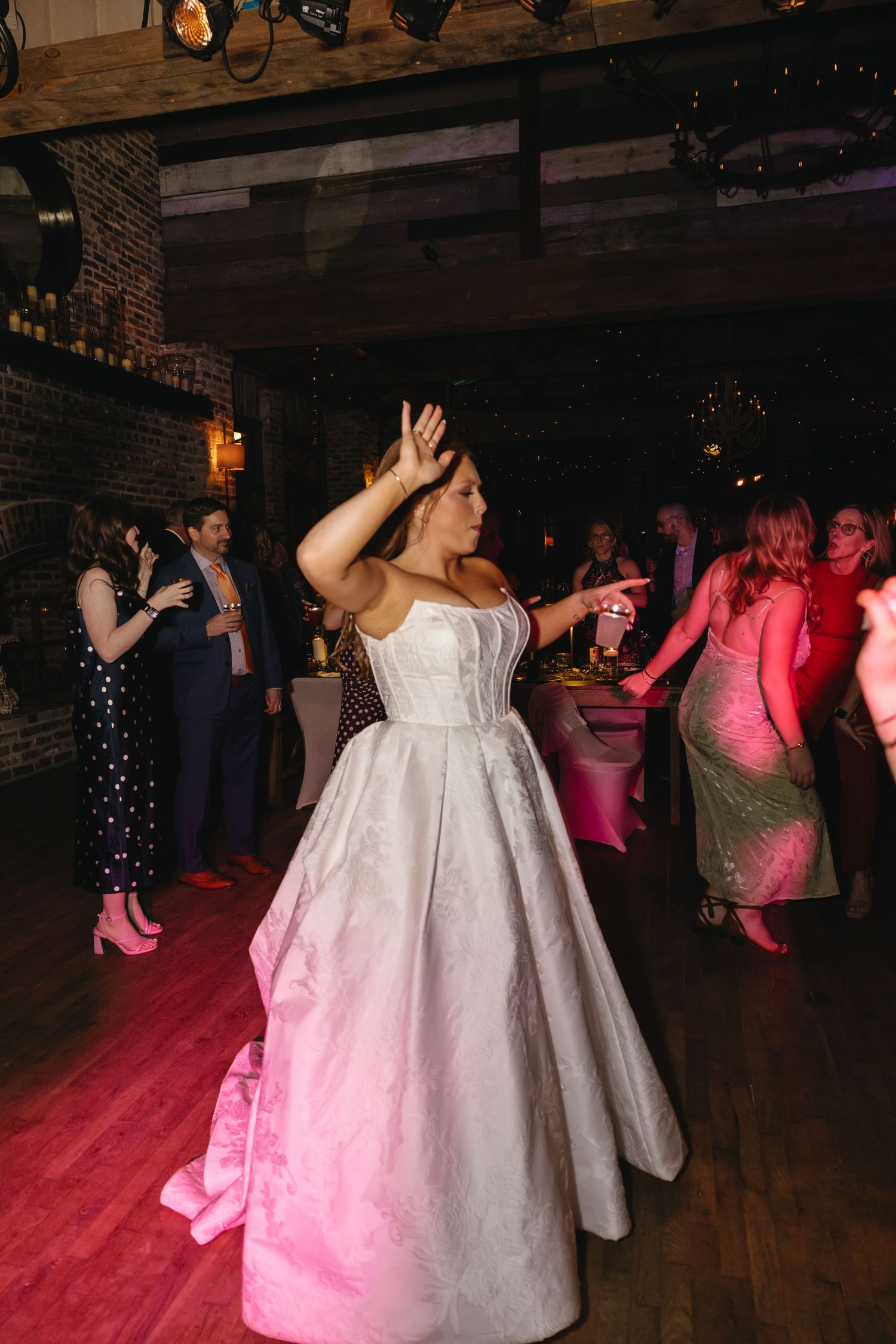 Bride in white ballgown dancing joyfully at wedding reception on dance floor with pink lighting.