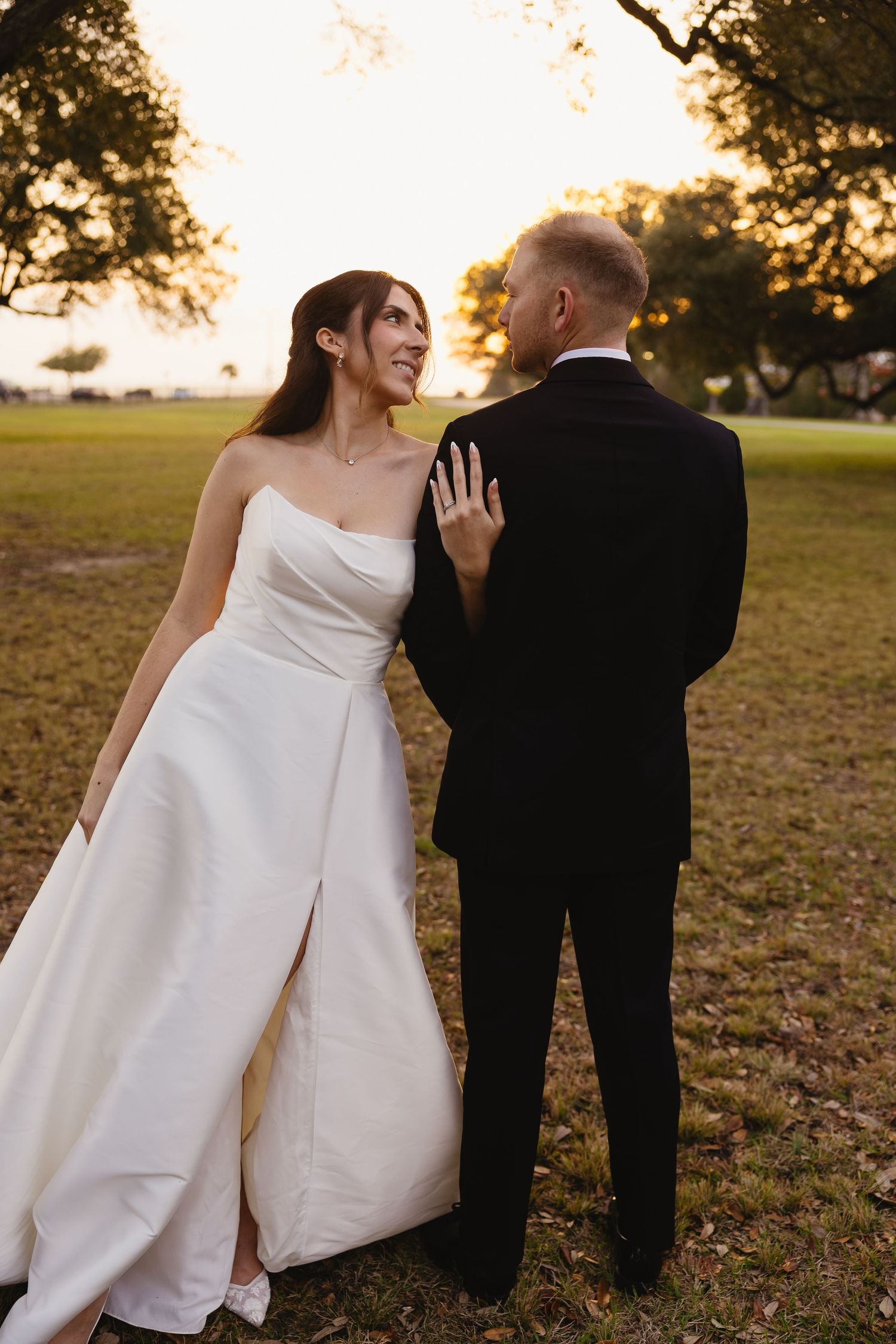 Bride in strapless white gown gazes at groom in black tuxedo during golden hour outdoor wedding portrait.