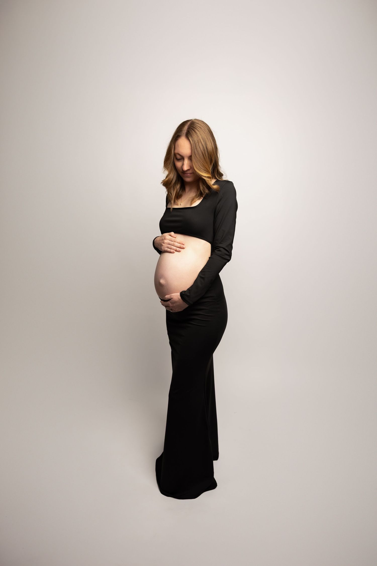 Pregnant woman in elegant black long-sleeve gown cradles baby bump in studio portrait.