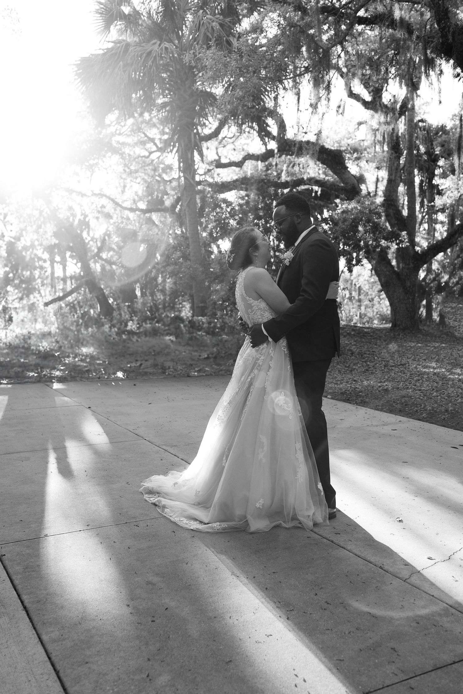 Black and white photo of bride and groom embracing under sunlit moss-draped trees.