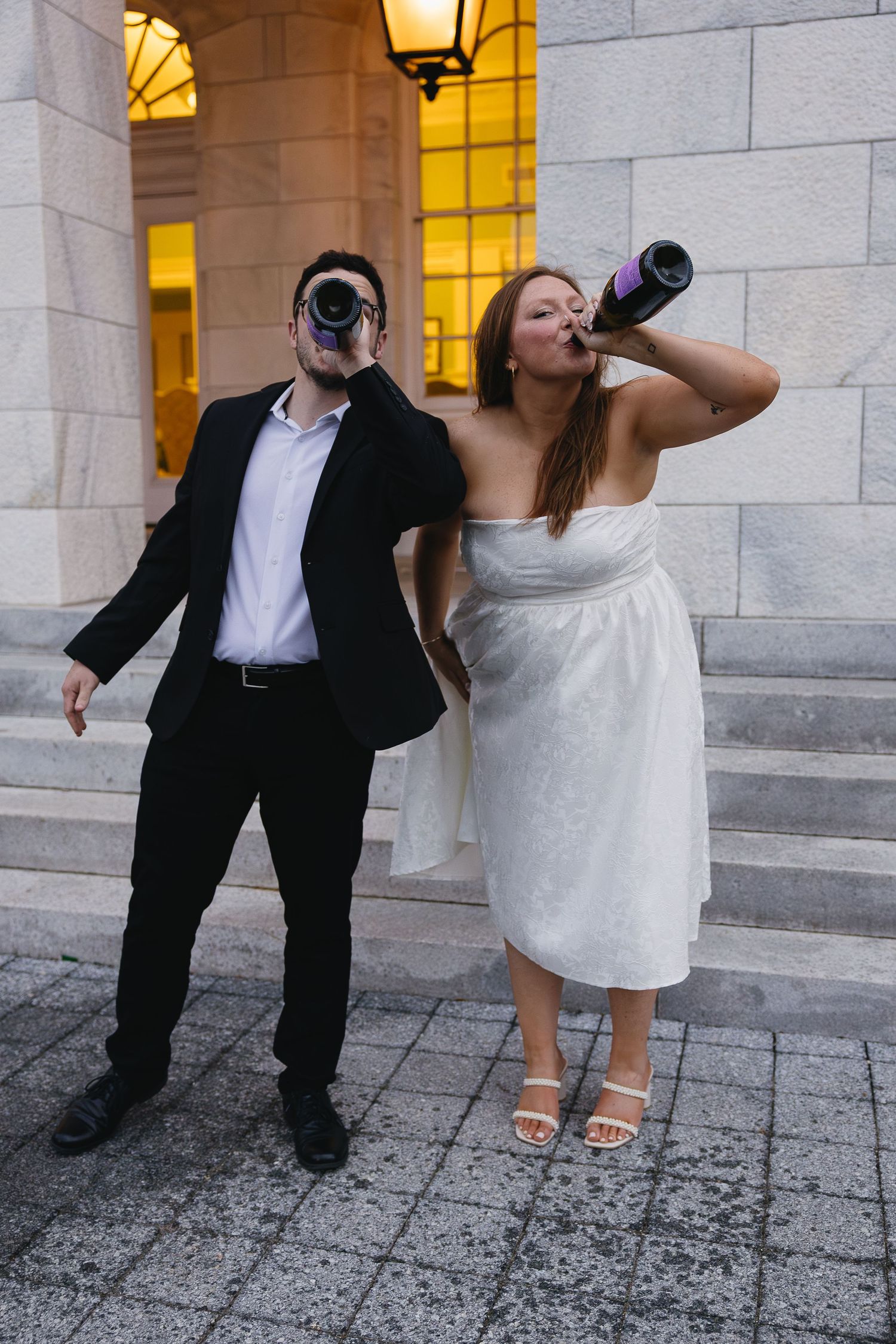 Playful newlyweds drinking champagne straight from bottles outside building after wedding celebration.