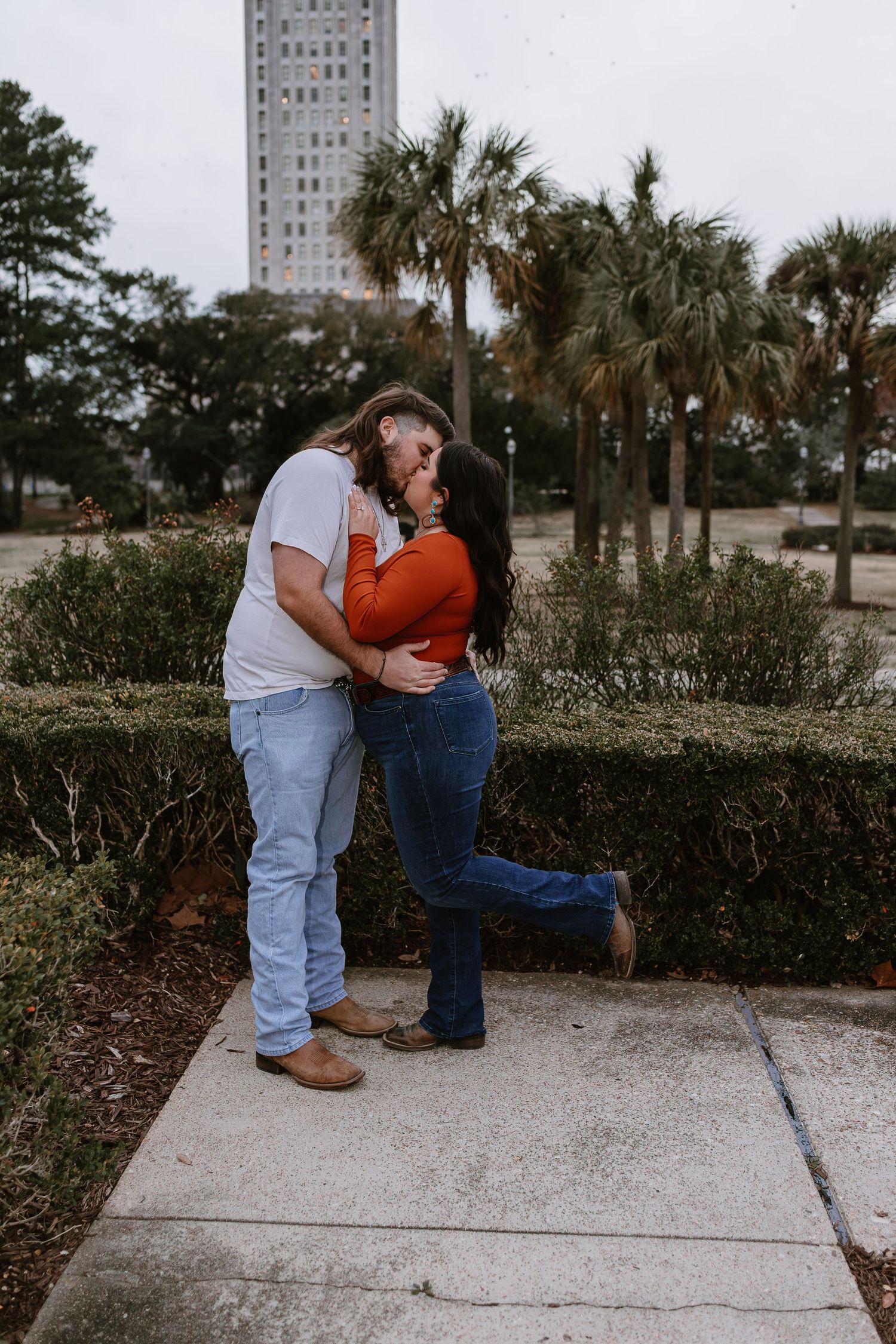Couple kissing near trimmed hedges with palm trees and a tall tower visible in the background.