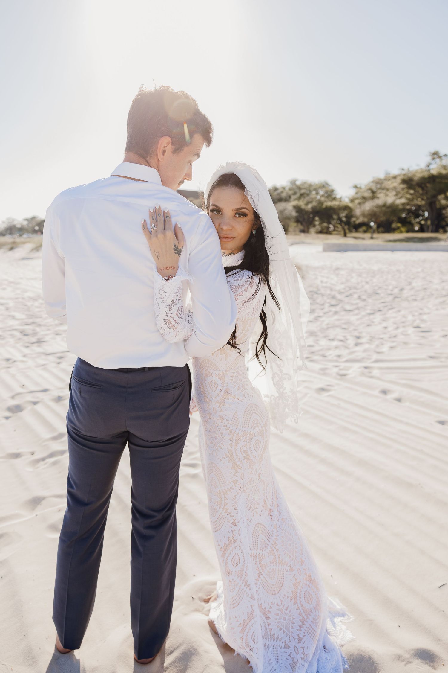 Tattooed bride in lace gown gazes back at camera while embracing groom on beach.