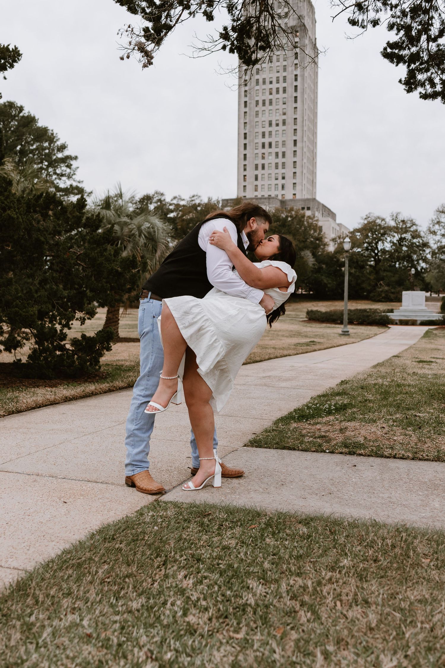 Groom dips bride in romantic kiss on pathway with Louisiana State Capitol building in background during engagement session.
