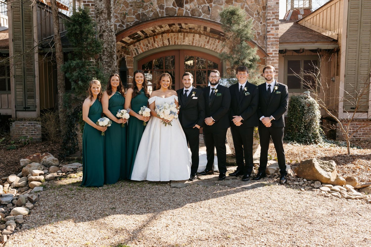 Wedding party in emerald green and black attire posing in front of rustic stone venue exterior.