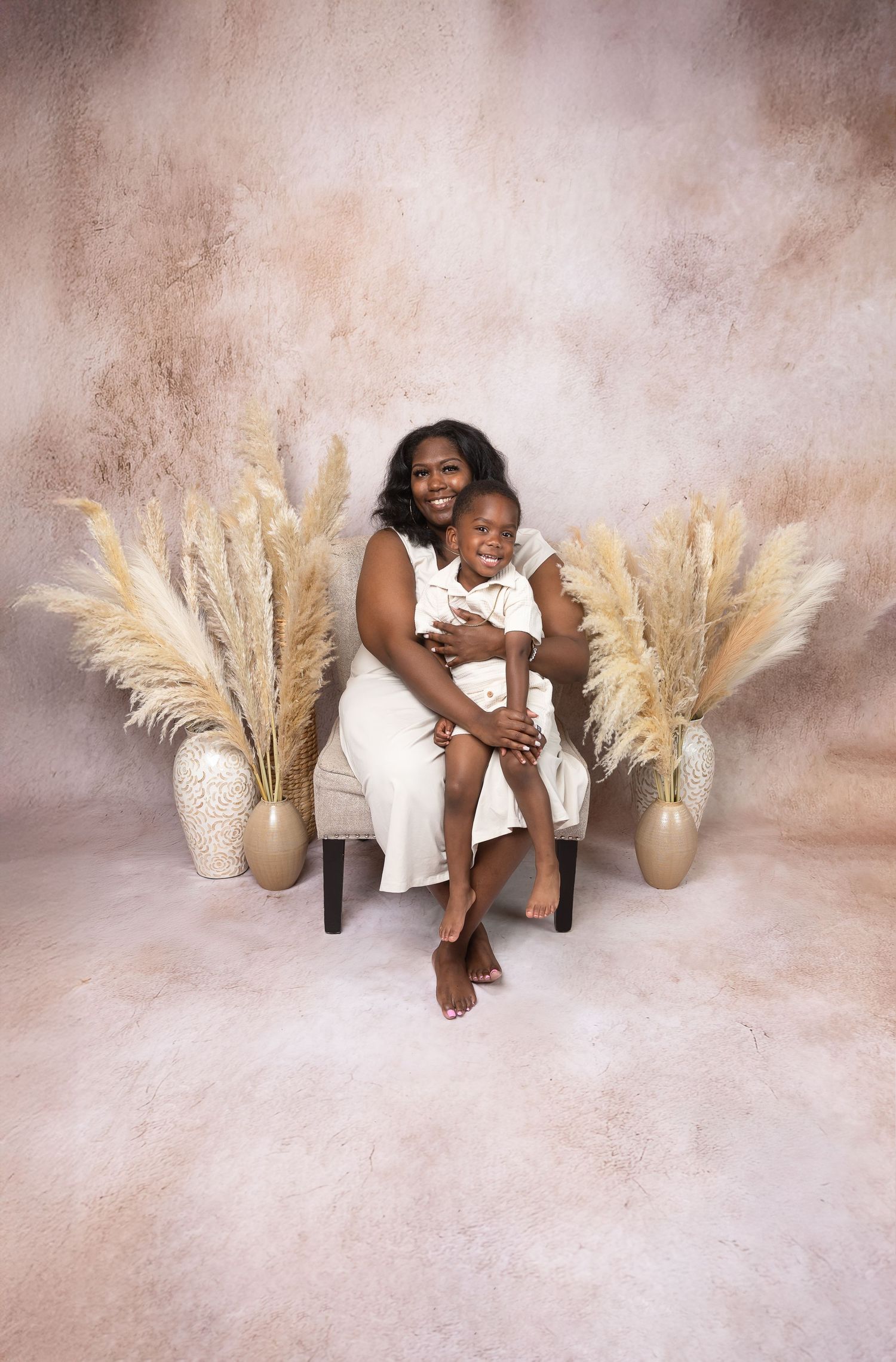 Mother in white dress sits on chair holding young child surrounded by pampas grass decor.