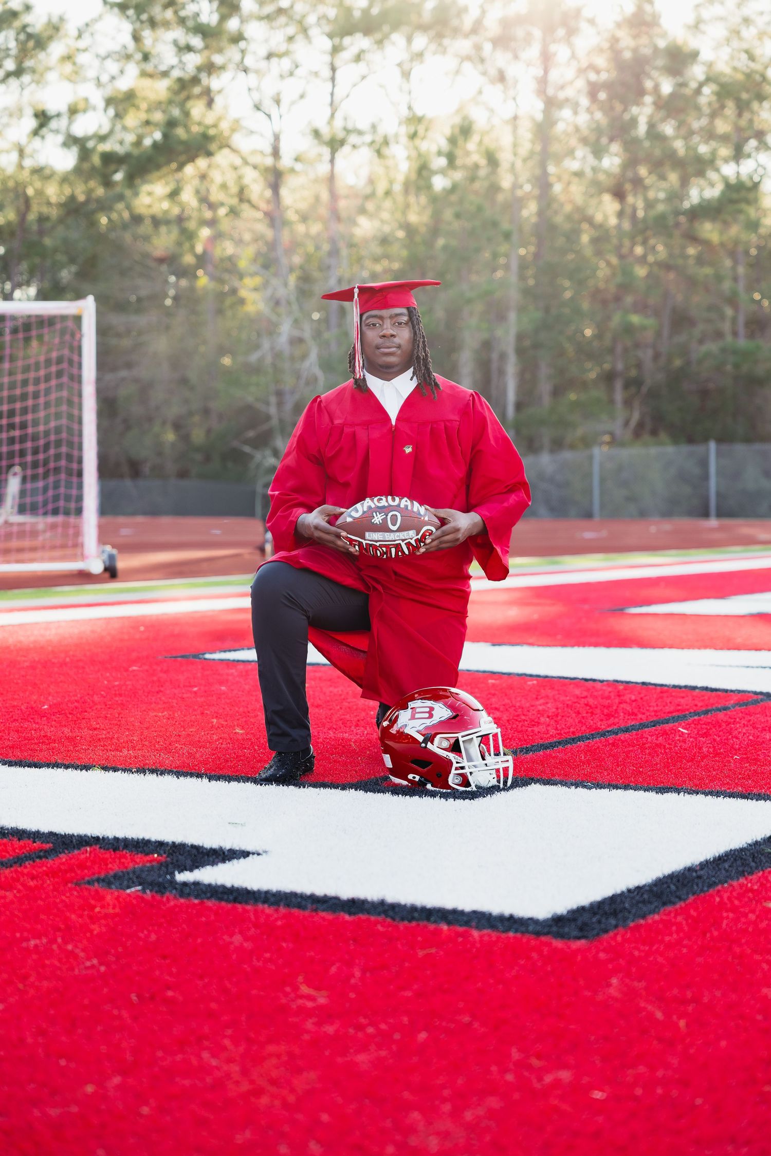 Graduate in red cap and gown kneeling on red turf field holding football with helmet beside him.