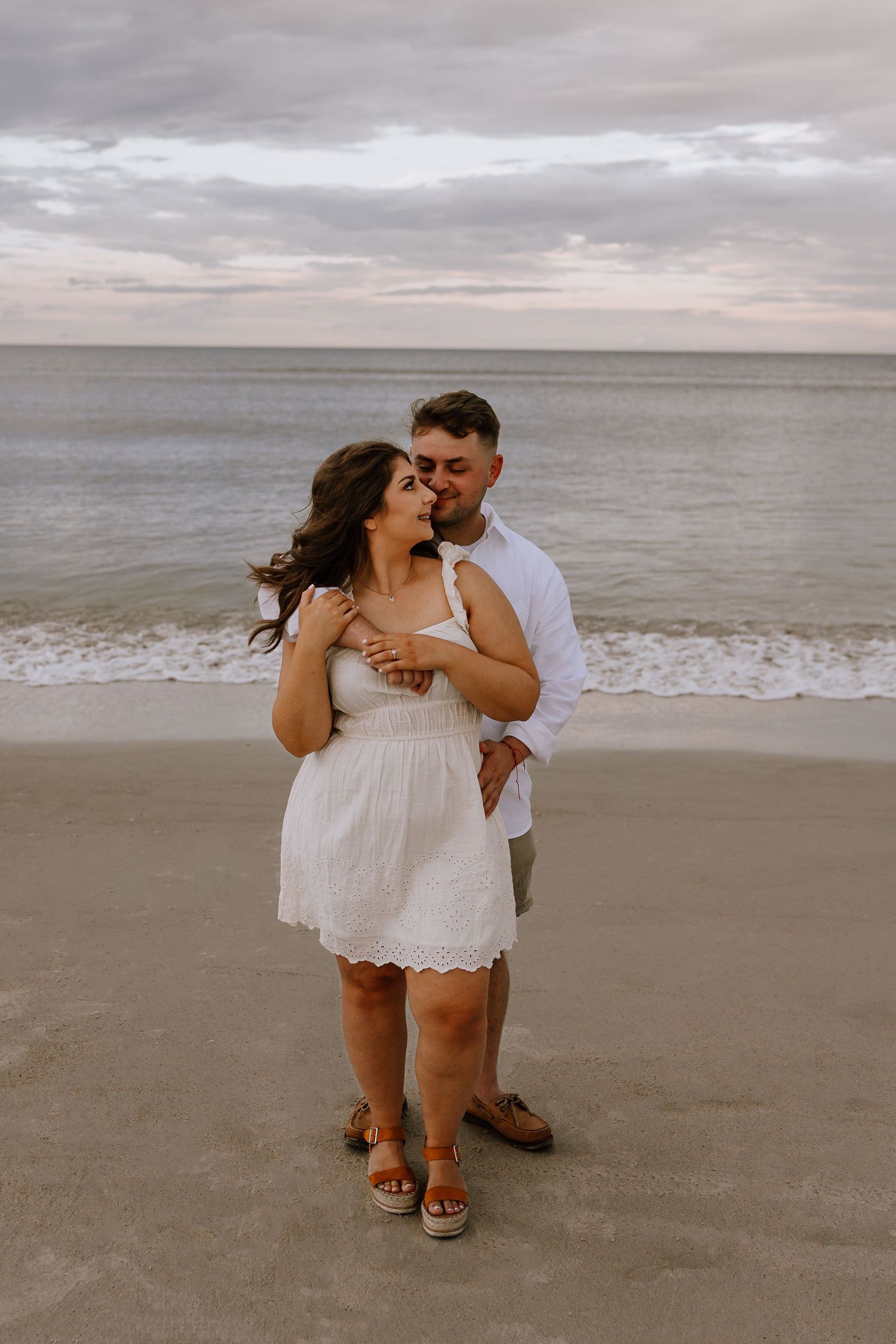 Romantic couple kissing on a sandy beach at dusk, woman in white dress and man in white shirt.