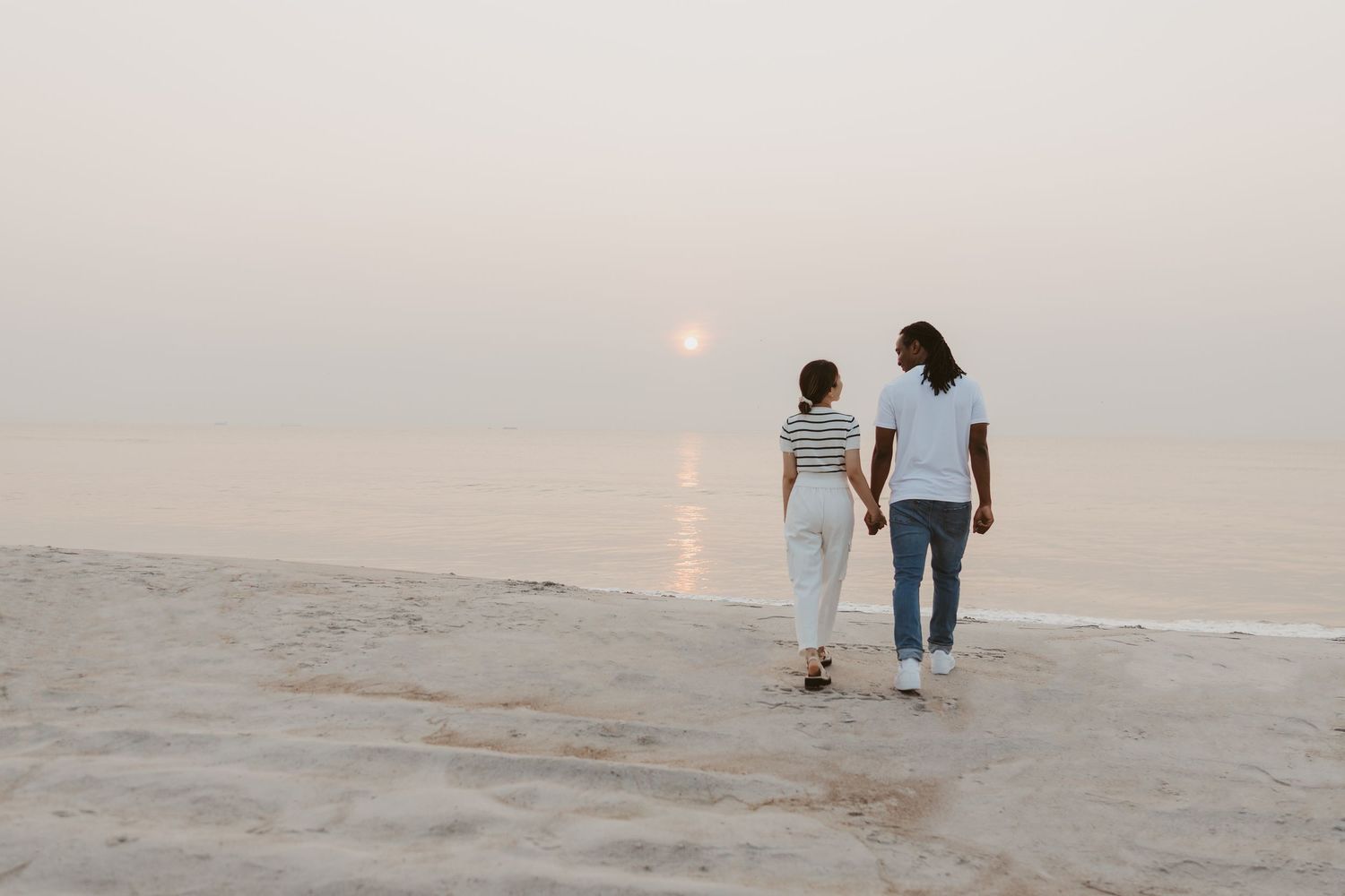 Couple holding hands walking along a sandy beach toward a glowing sunset.