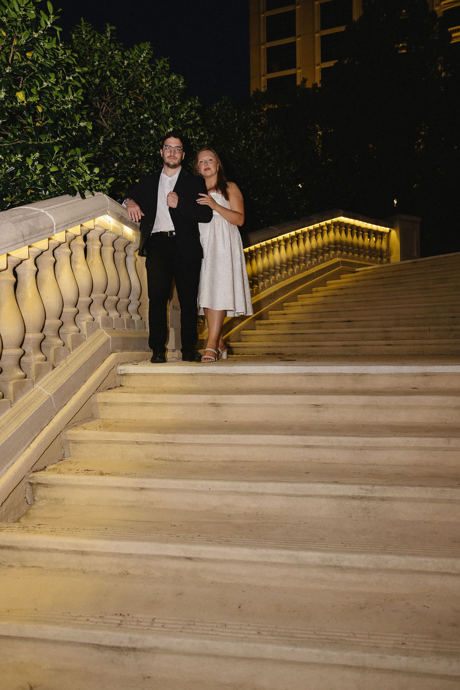 Smiling newlyweds descending illuminated grand staircase together at night after wedding ceremony.