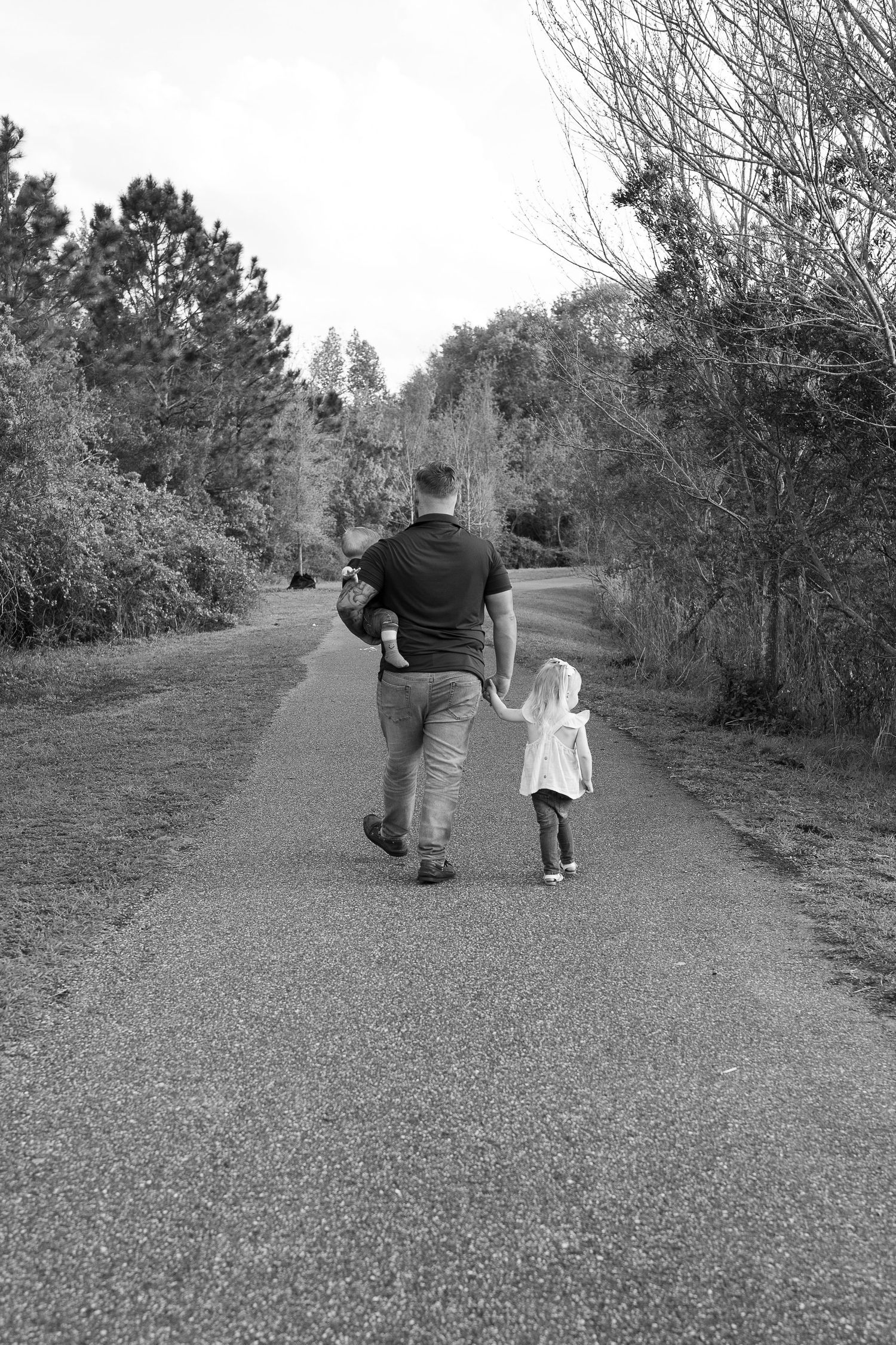 Black and white photo of a father walking hand-in-hand with his young child down a tree-lined path.
