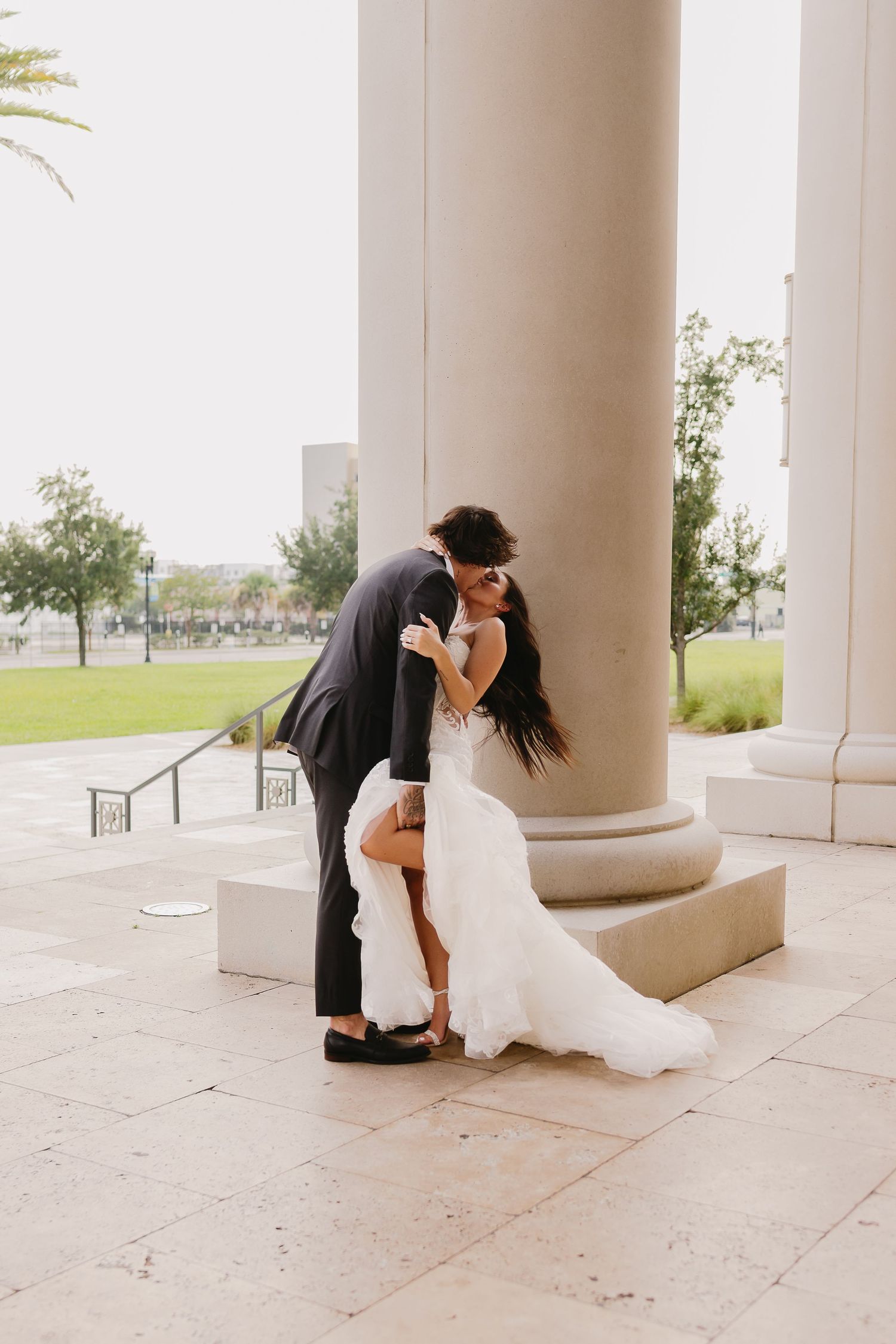 Groom dips bride in white mermaid gown beside stone column in romantic wedding pose.
