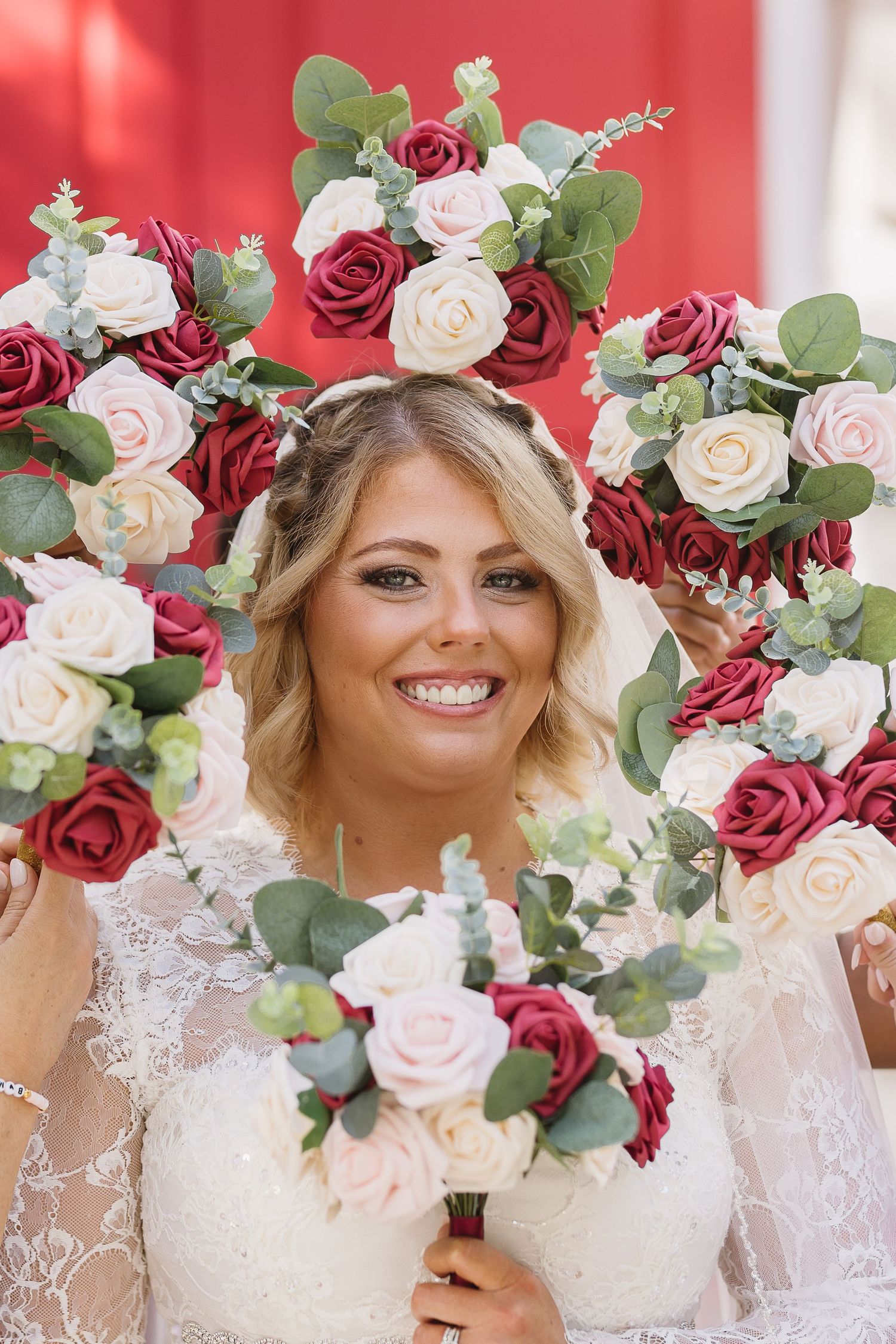 Smiling bride framed by burgundy and white rose bouquets on her wedding day.