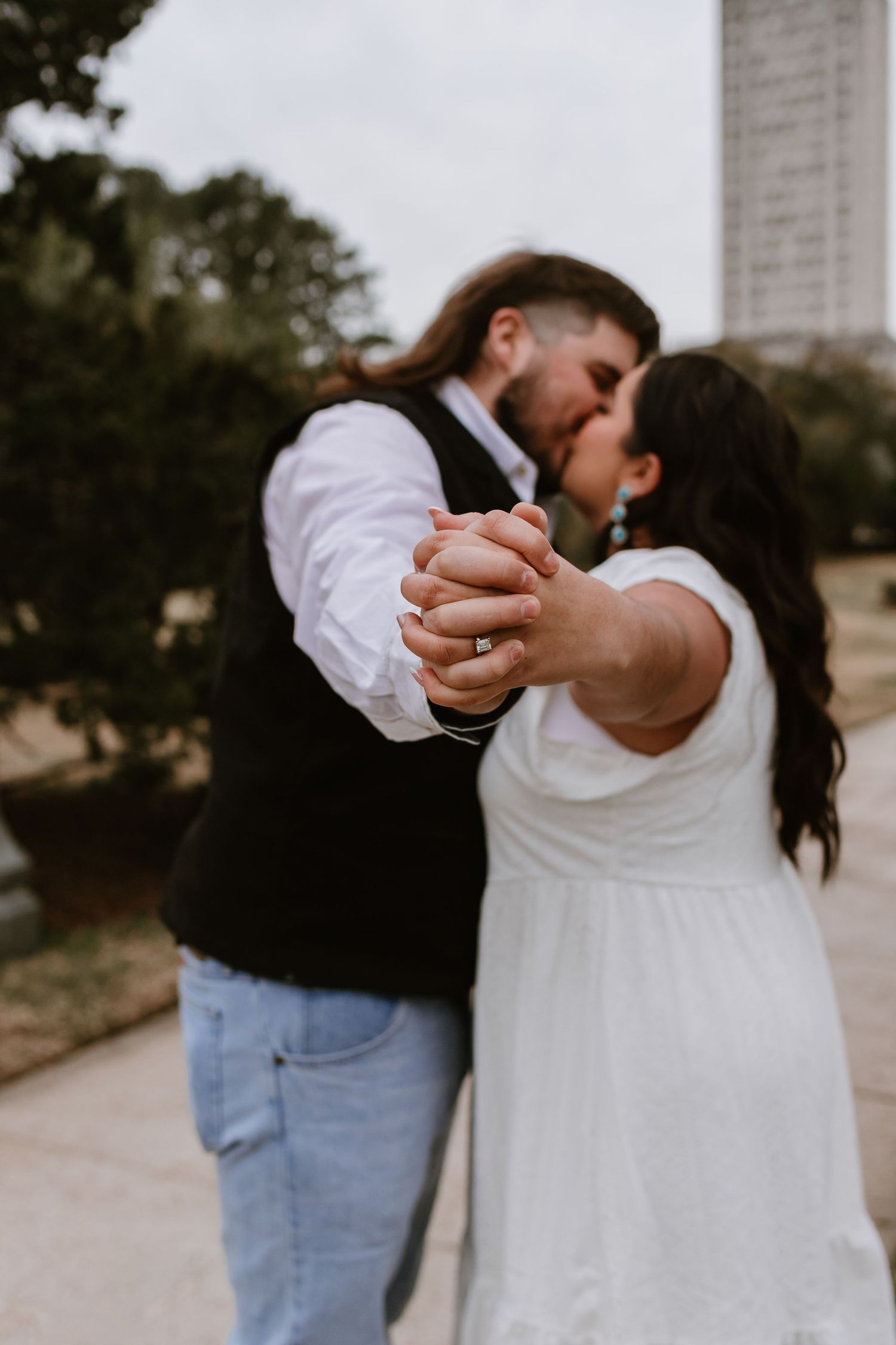Couple shares tender kiss while holding hands, diamond engagement ring visible, with urban backdrop during outdoor photo session.