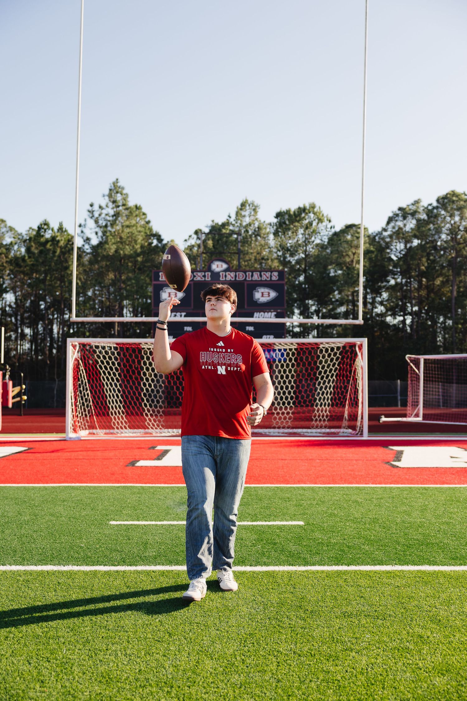 Young man in red Nebraska shirt tossing football on red turf football field.