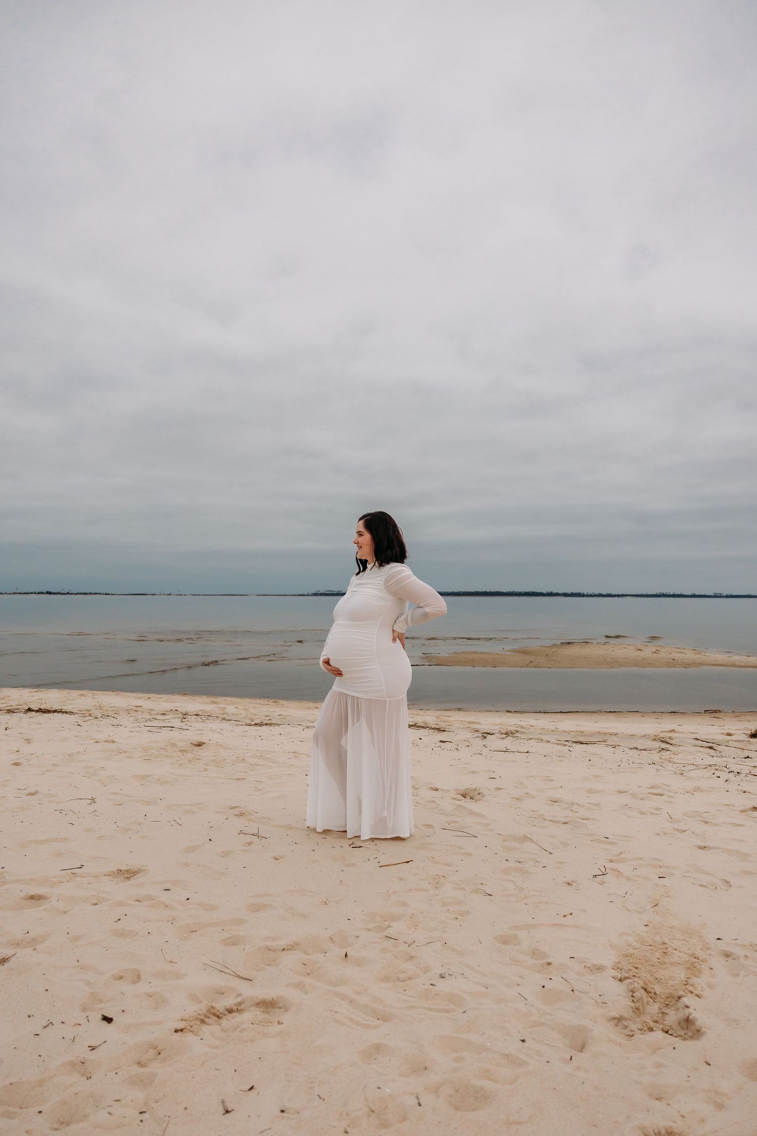 Expecting mother in flowing white gown posing on serene sandy beach.