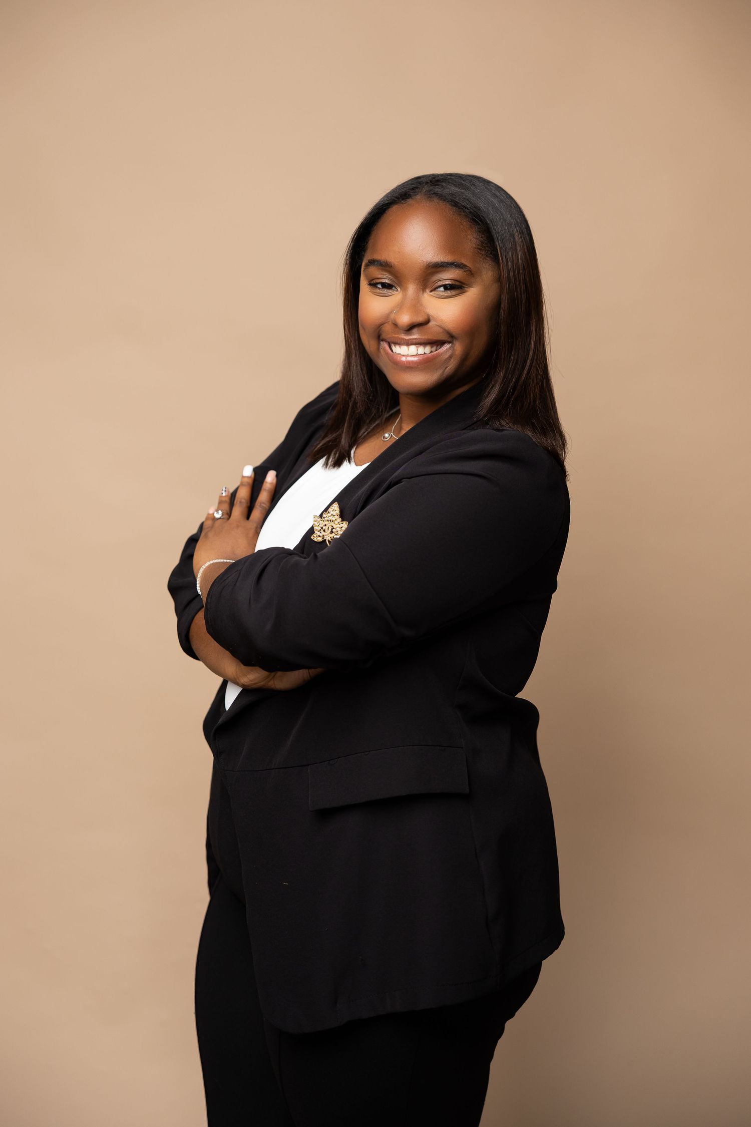 A confident young Black woman in a black blazer smiles with arms crossed against a beige background.