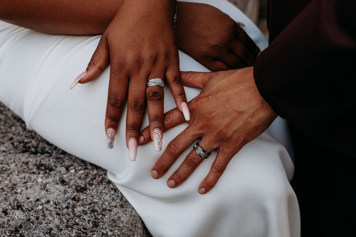 Close-up of bride and groom's hands showing wedding rings on white dress.