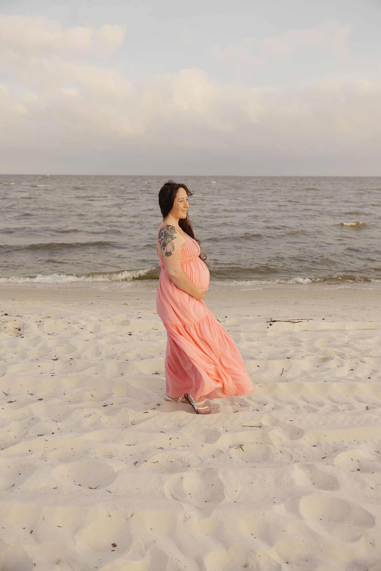 Serene pregnant woman in flowing pink dress standing sideways on beach, ocean in background.
