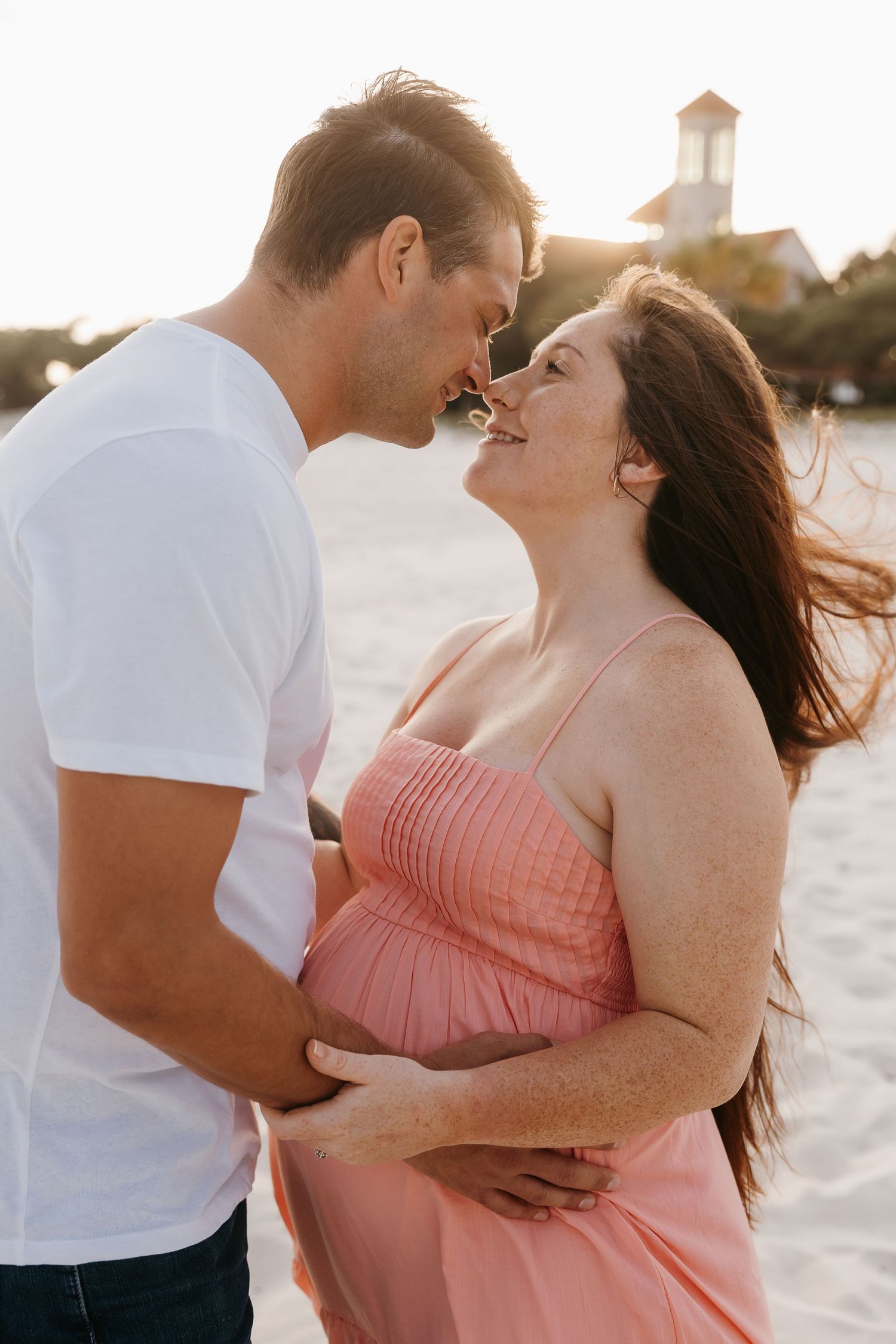 Couple sharing an intimate moment at sunset beach, pregnant woman in pink dress smiling at partner.