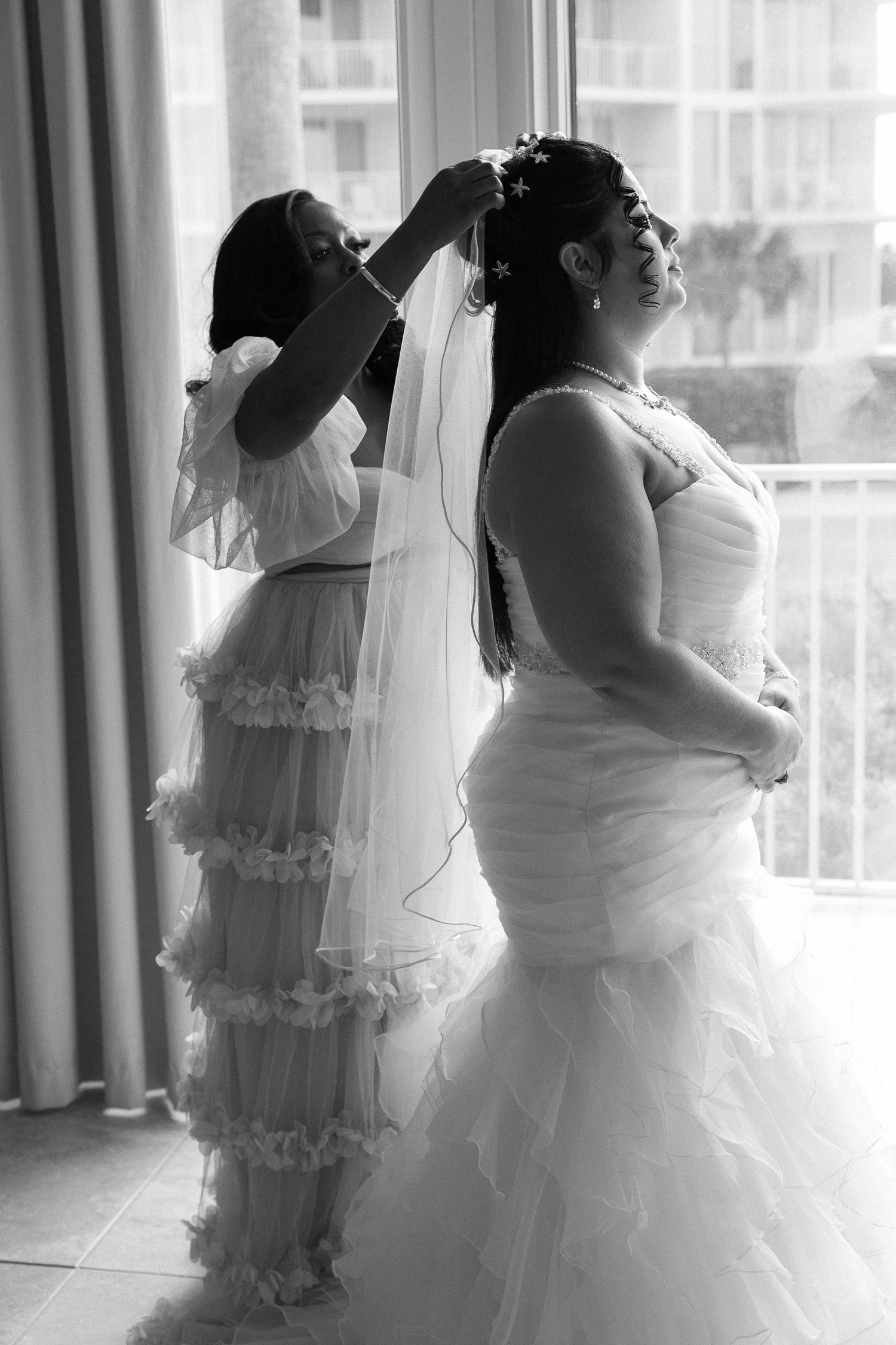 Black and white photo of bridesmaid pinning veil into brides hair near hotel window.