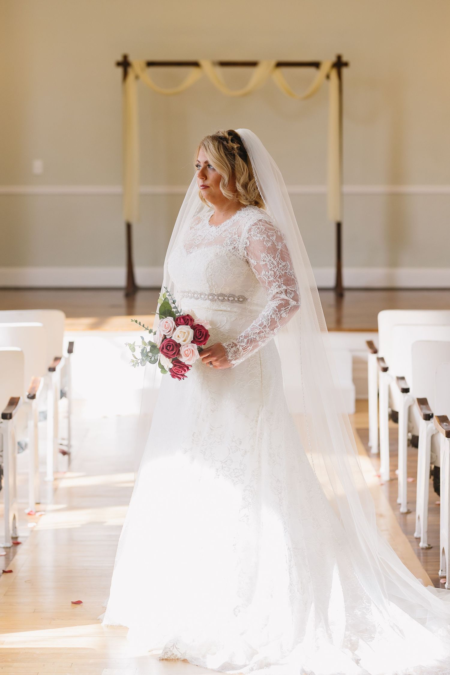 Bride in lace long-sleeve gown and veil holding bouquet in sunlit wedding chapel.