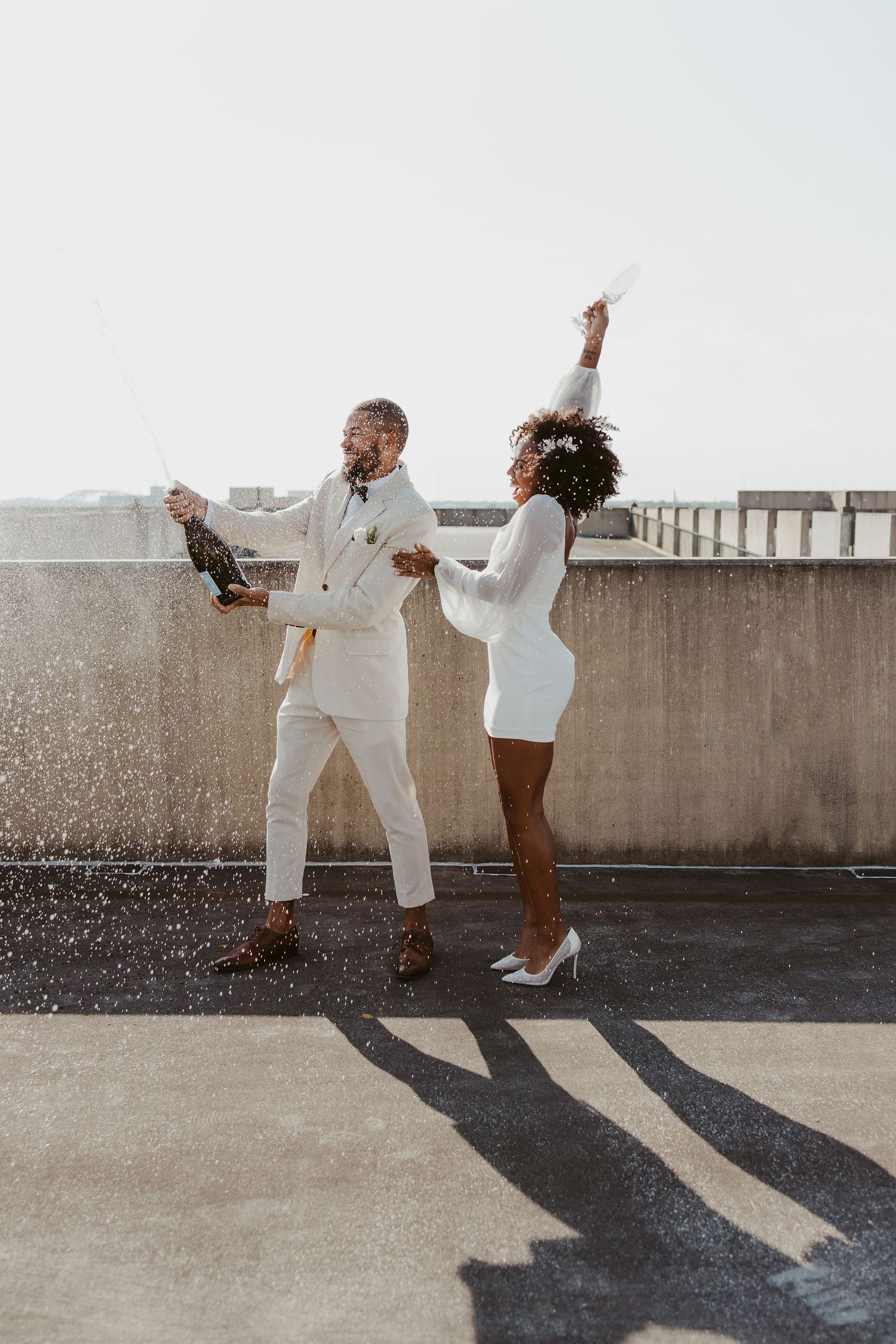 Joyful newlywed couple popping champagne on rooftop in matching white wedding outfits.
