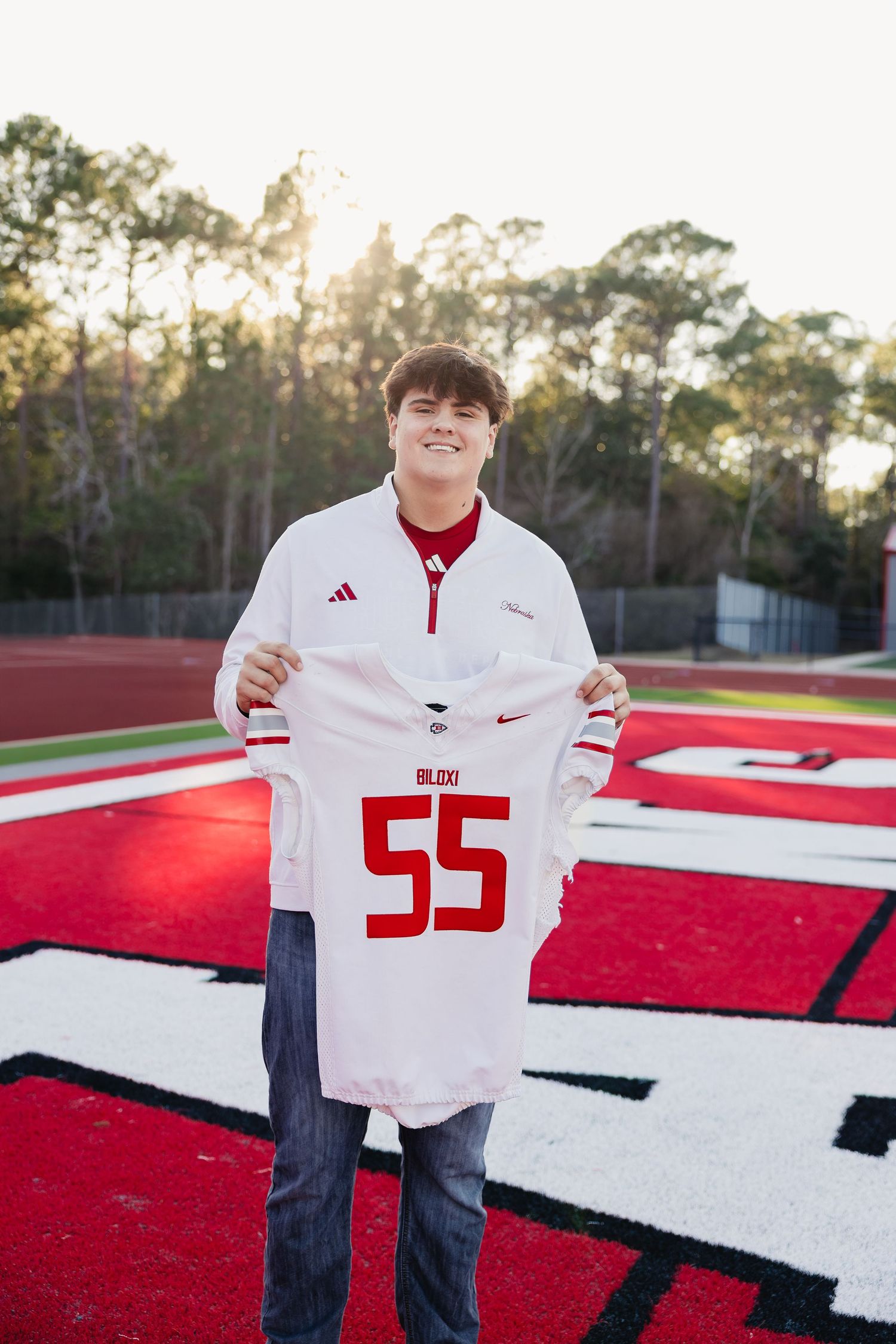 Smiling teen holding white Biloxi number 55 football jersey on red turf field.