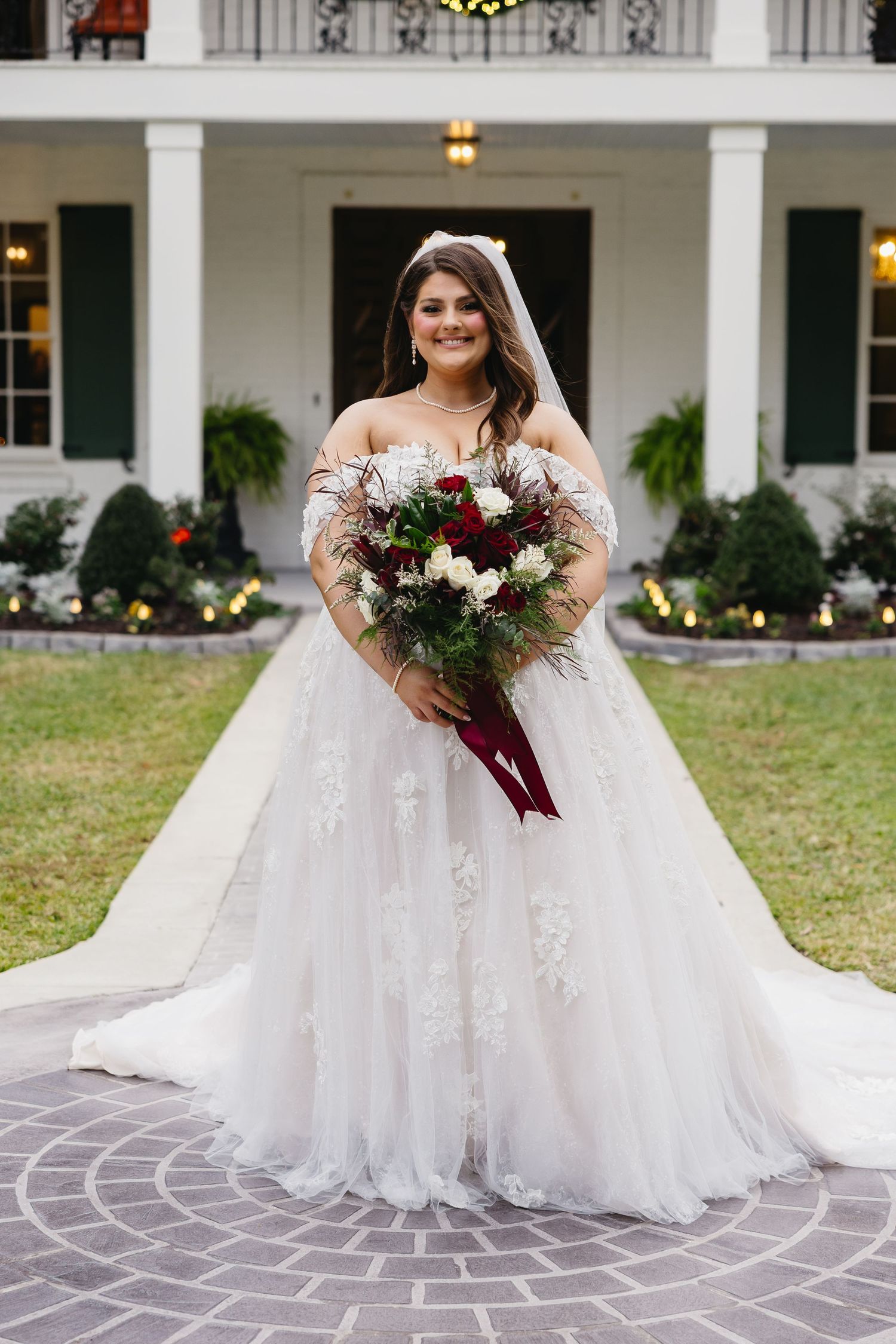 Bride in off-shoulder lace ballgown holding red and white bouquet stands before a white Southern mansion.