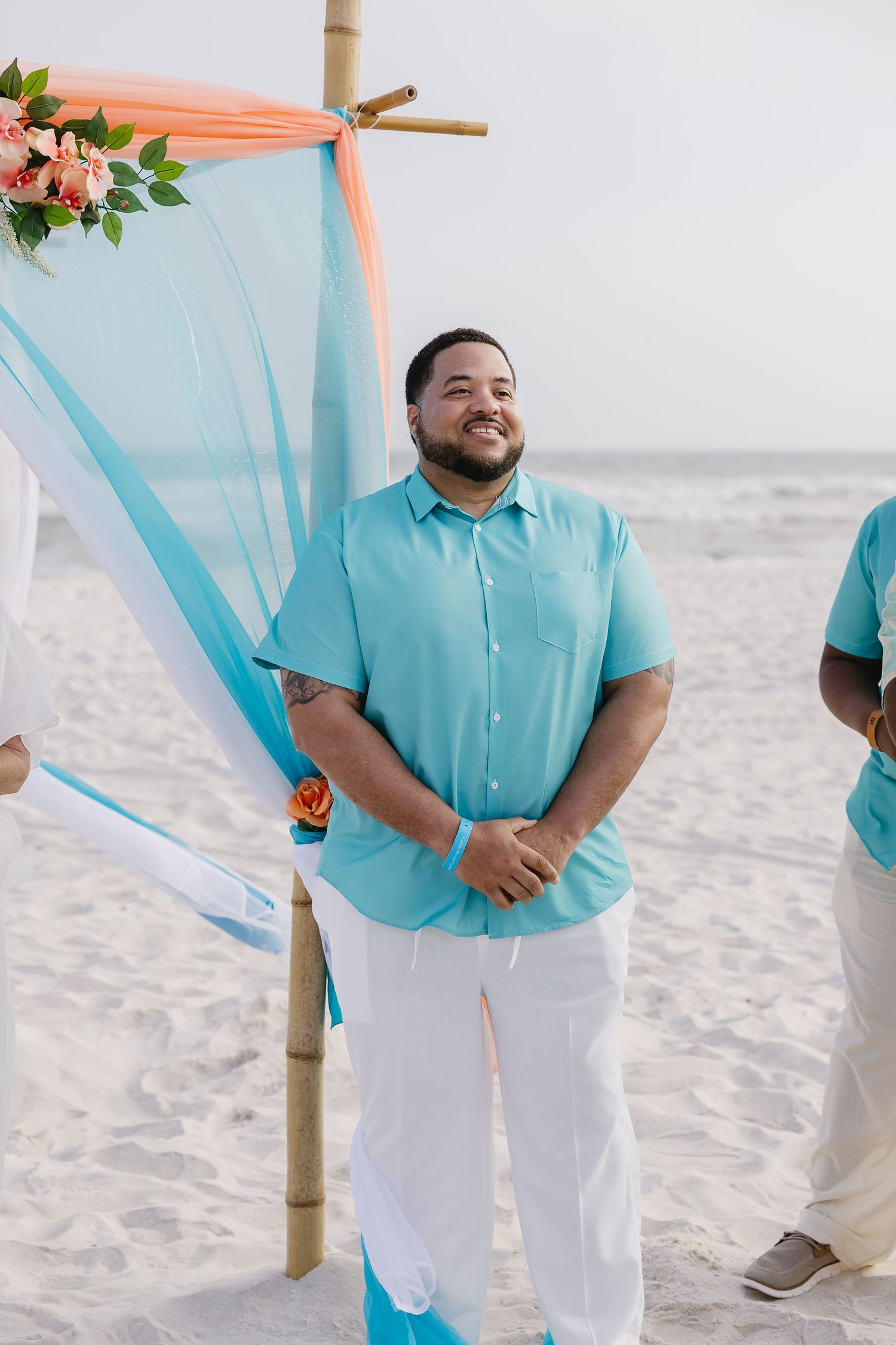 Smiling groom in turquoise shirt stands at bamboo beach wedding arch decorated with flowers.