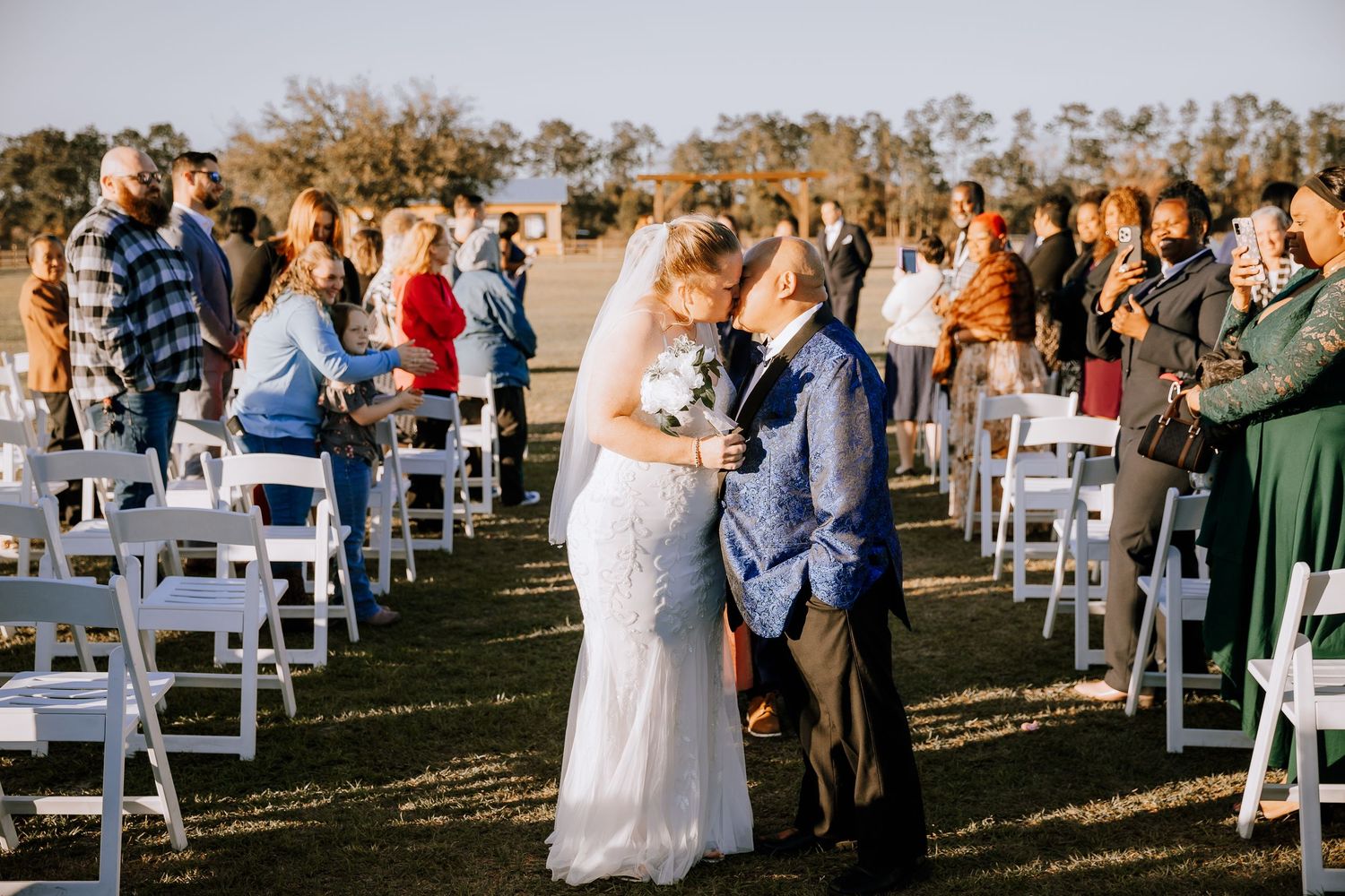 Bride and groom kissing at the end of the aisle after outdoor wedding ceremony with guests applauding.