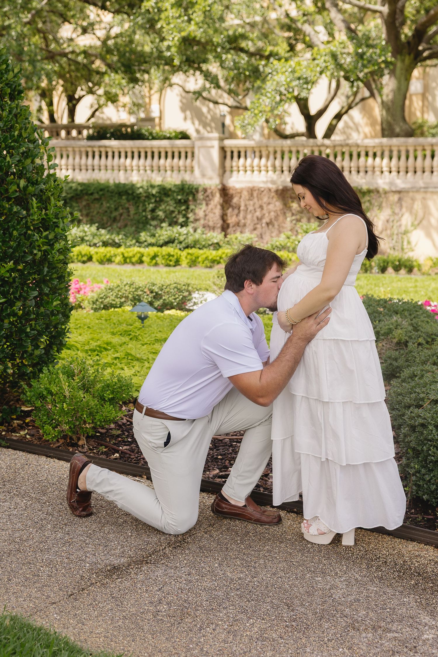 Man kneeling to kiss pregnant woman's belly in a lush garden setting, both dressed in white.