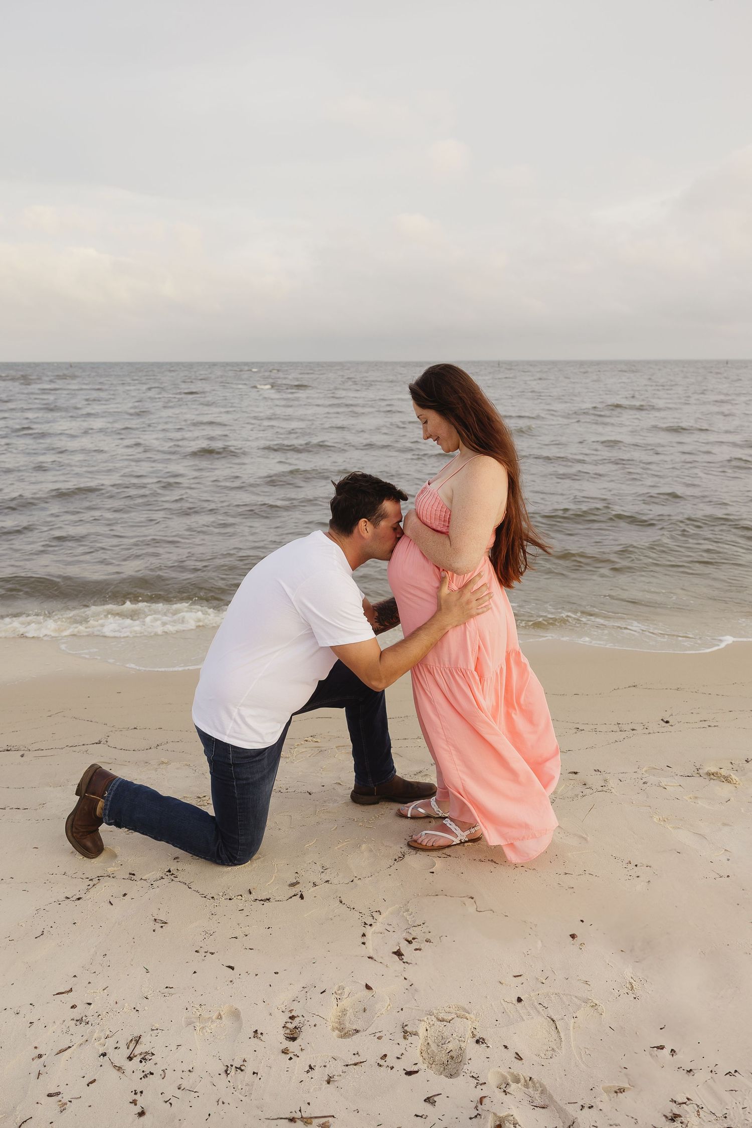 Man kneeling on beach kissing pregnant partners belly, tender maternity photo at seaside.