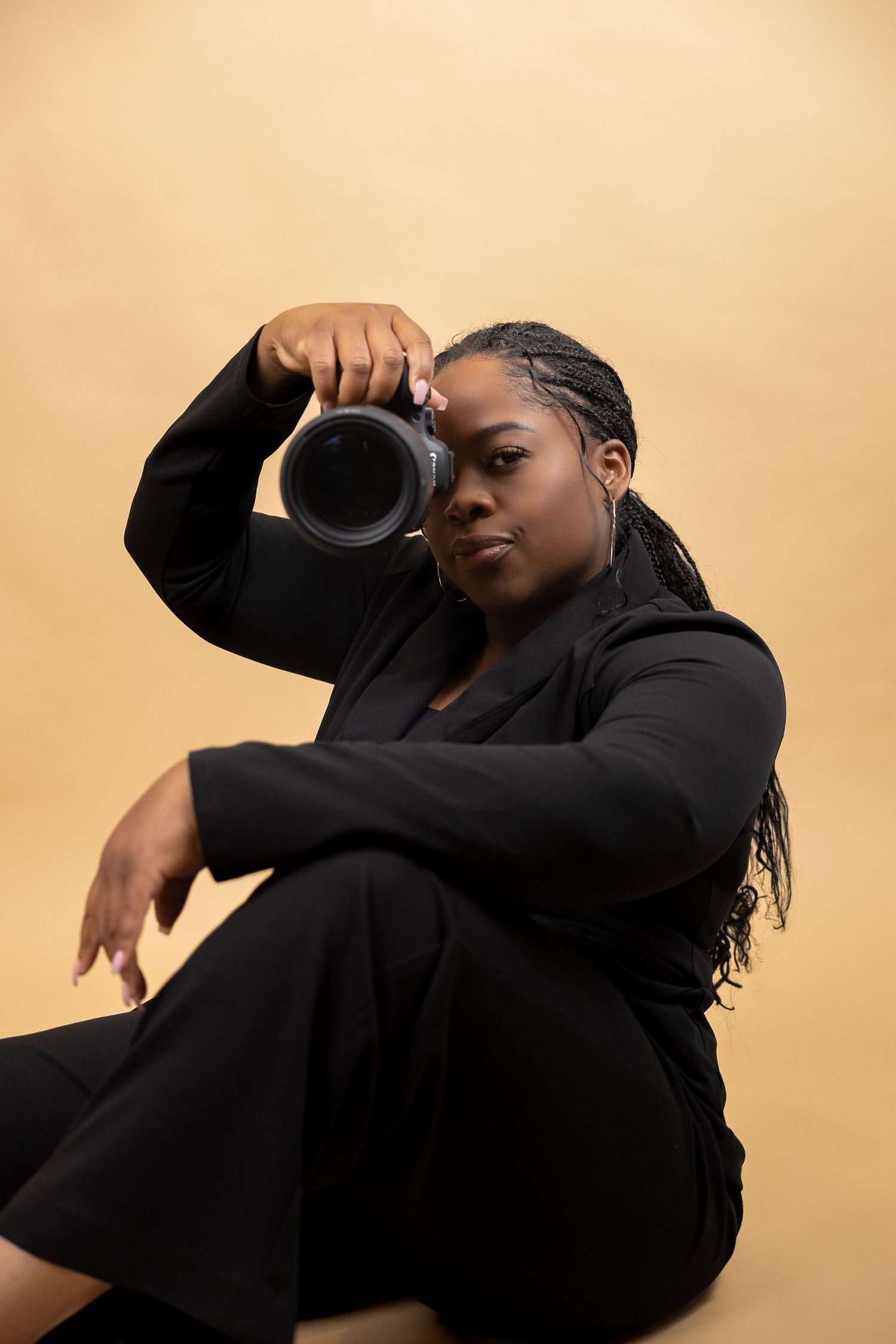Female photographer in black outfit holding camera while seated against a warm beige background.