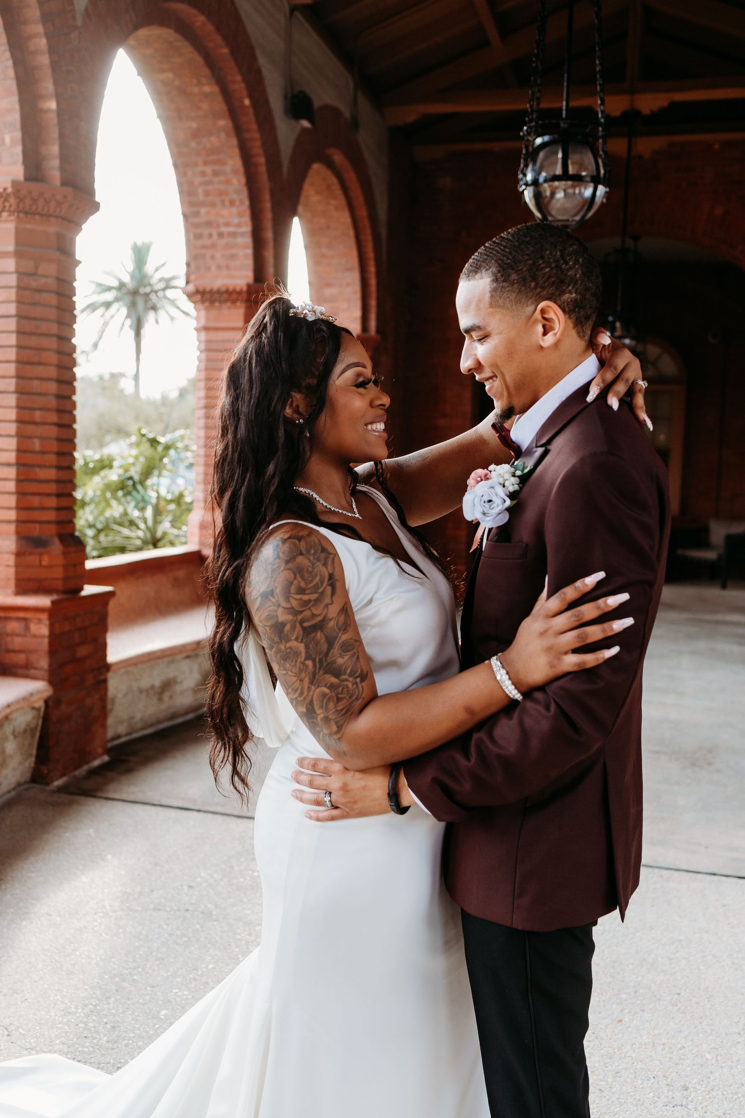 Bride with tattoo sleeve and groom in burgundy suit embracing and smiling.