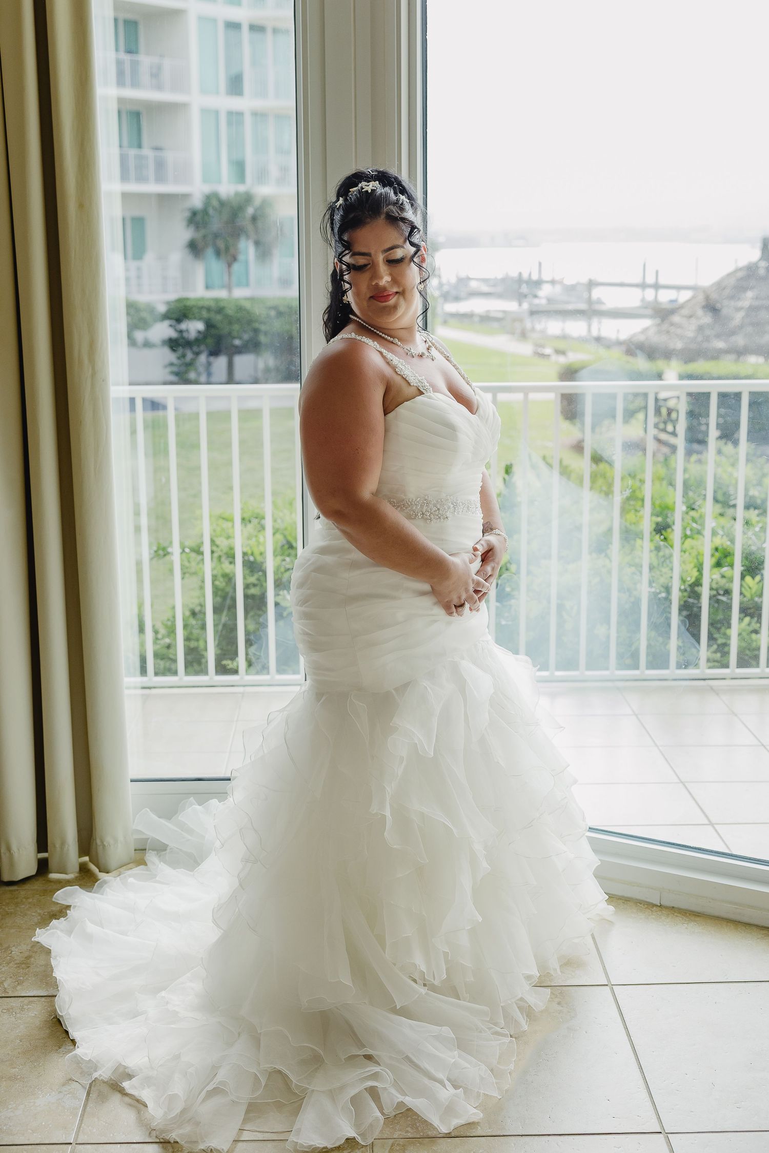 Bride in strapless mermaid wedding gown with ruffled train standing by window with garden view.