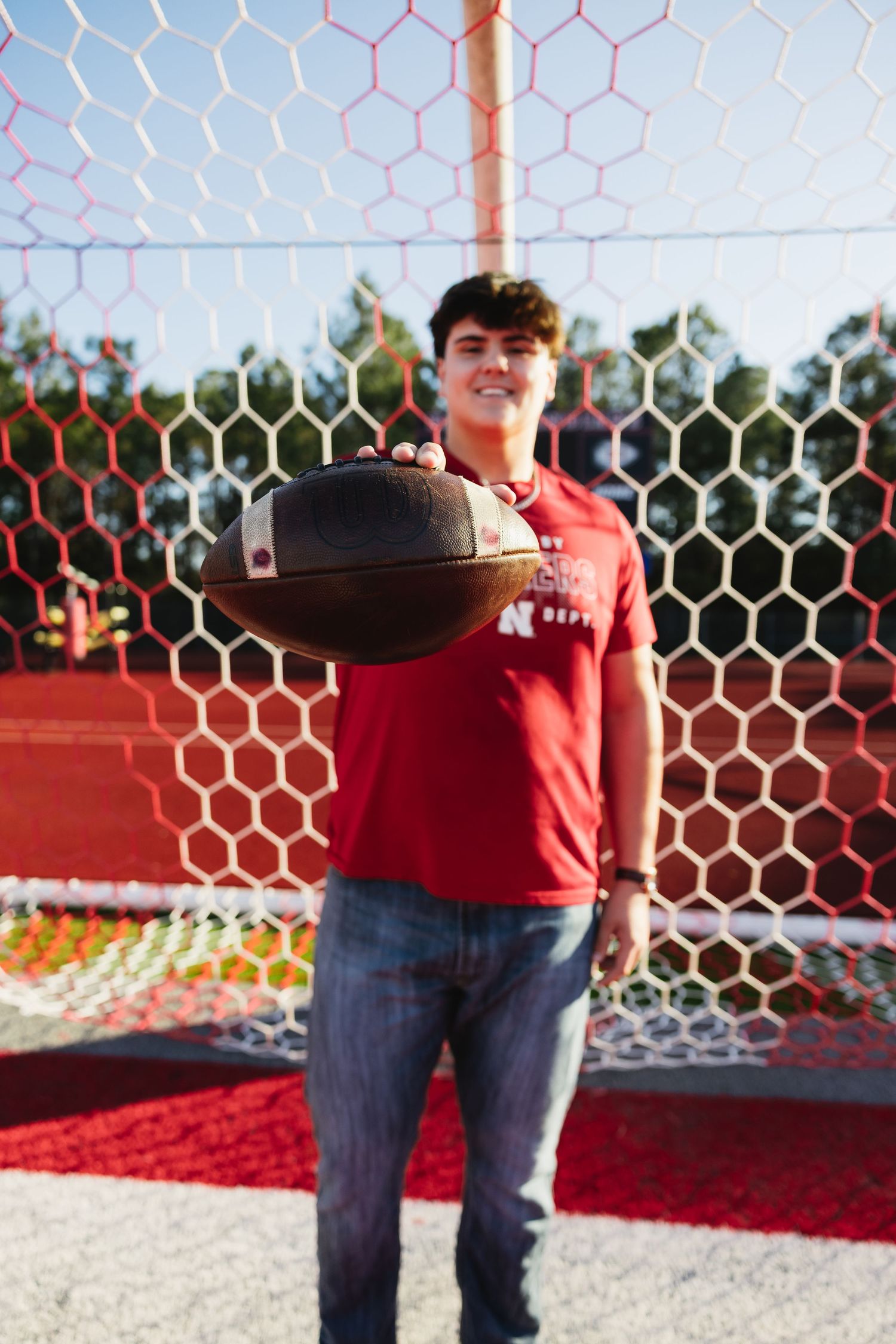 Smiling teen boy holding football in front of red goal net on athletic field.