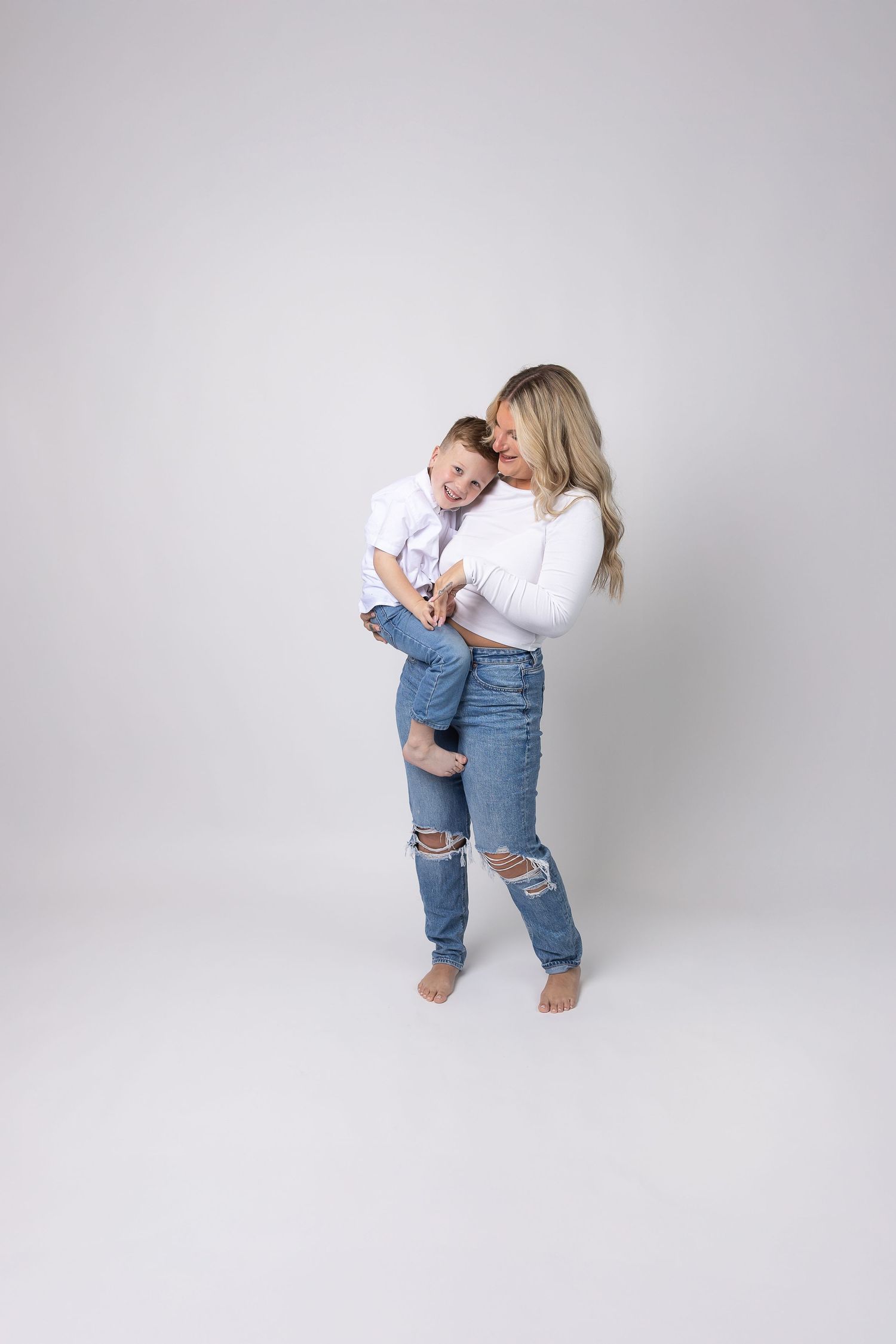 A mother in ripped jeans and white top holds her laughing toddler in a bright studio setting.