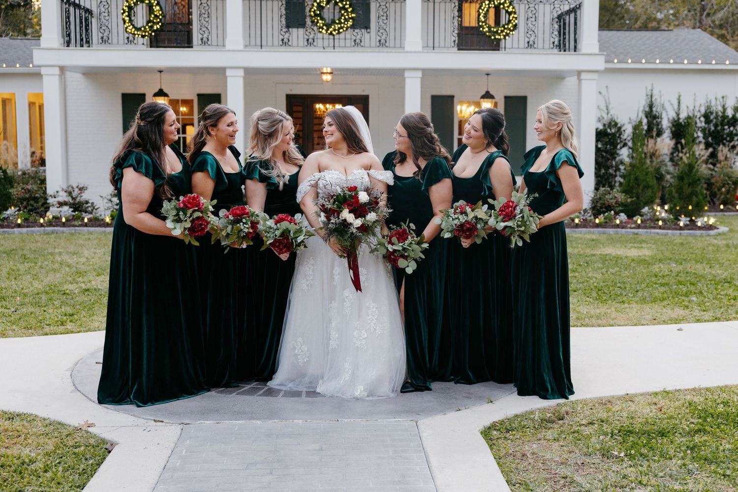 Bride with bridesmaids in emerald green velvet dresses holding red bouquets before white mansion.