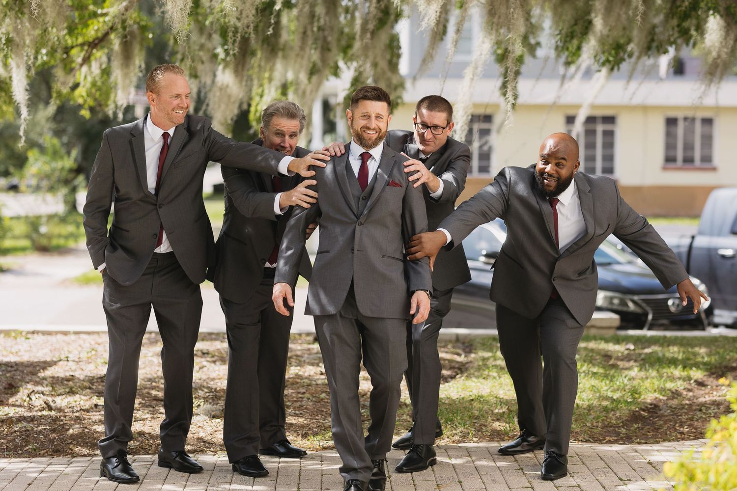 Groom and groomsmen in gray suits celebrating outdoors under Spanish moss trees.
