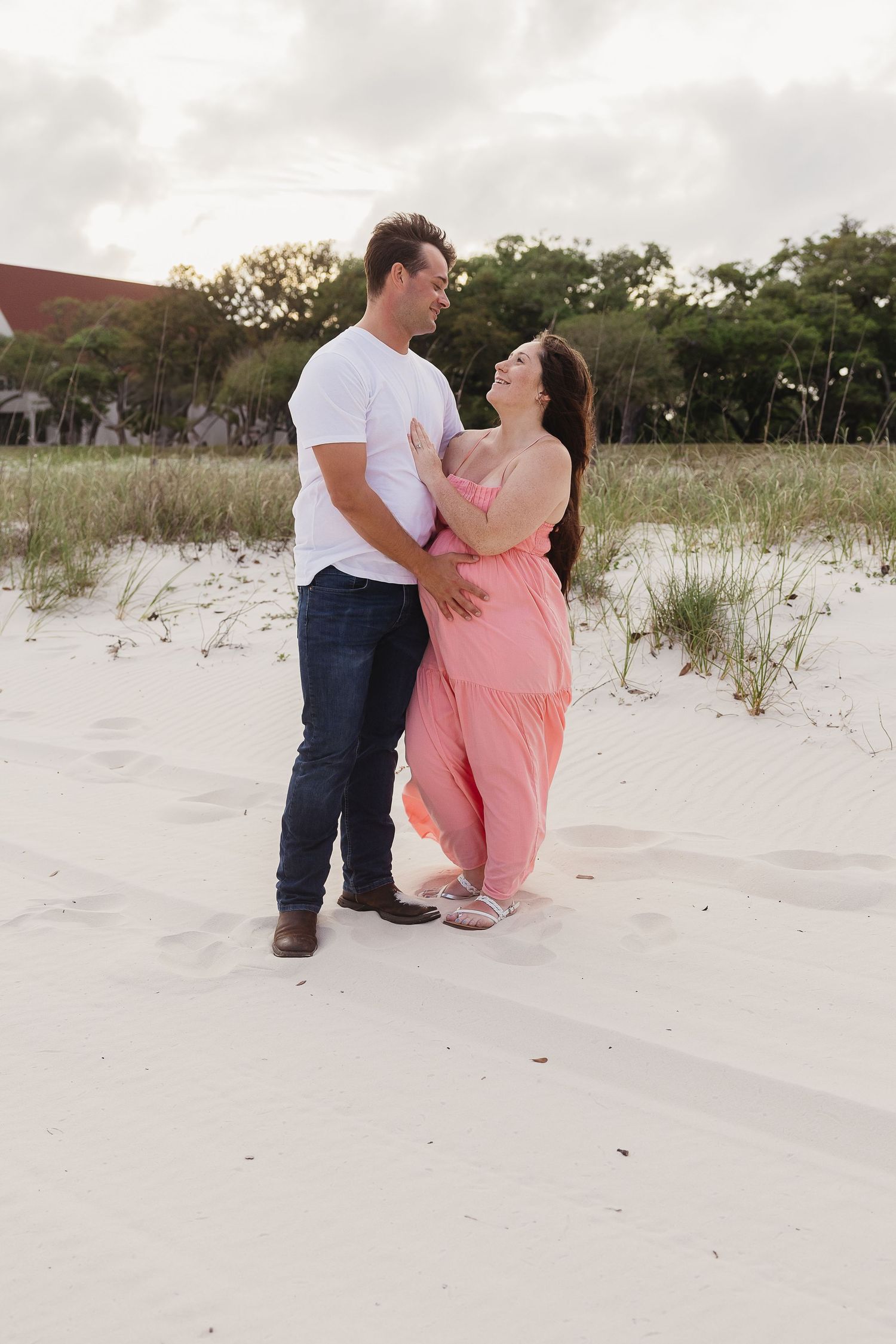 Happy expecting couple embracing on white sand dunes, woman in pink maternity dress laughing.