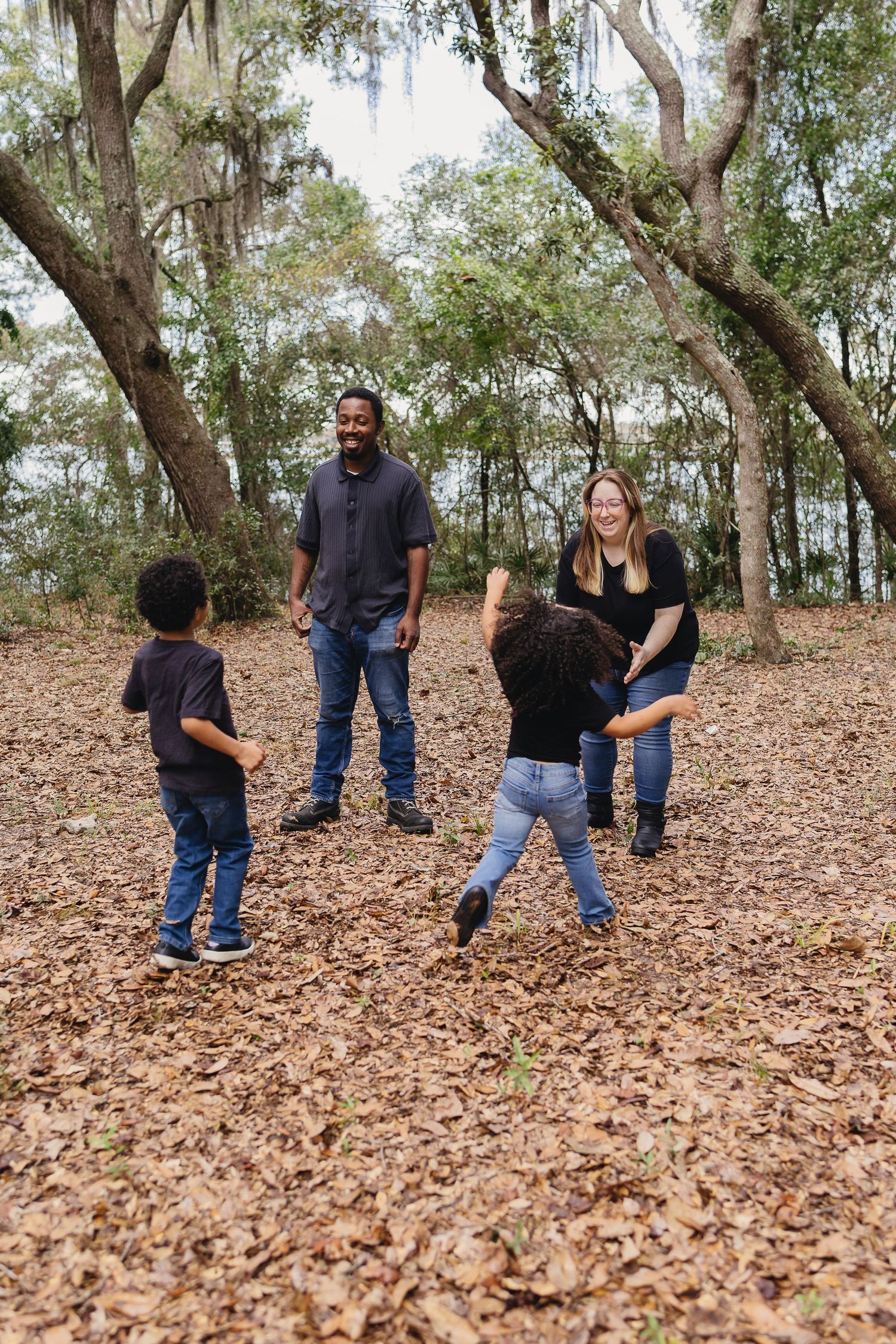A family of four playing and laughing together on a leaf-covered ground in a wooded park.