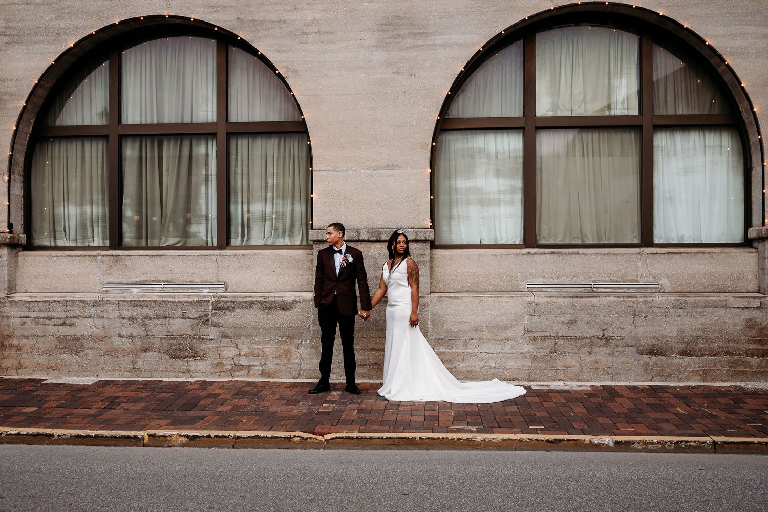 Bride and groom standing apart holding hands outside a stone building wall.