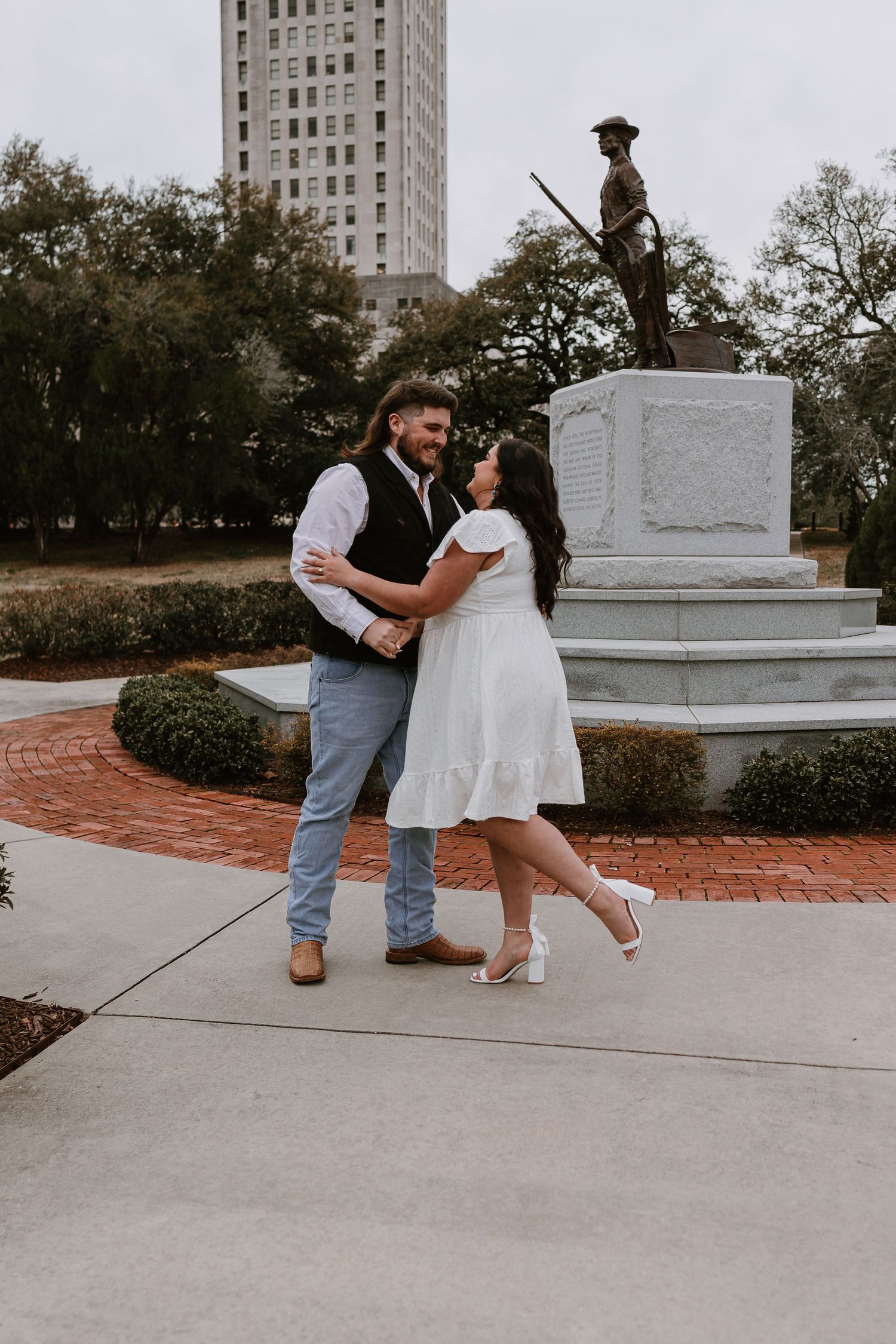 Couple dances and laughs together near bronze soldier statue with Louisiana State Capitol tower in background.