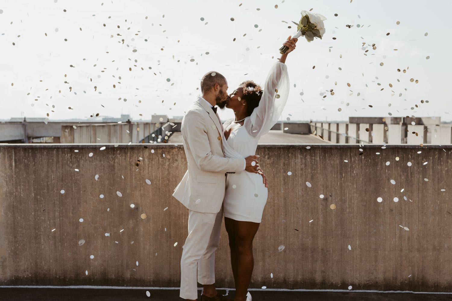 Interracial couple kissing on rooftop surrounded by falling white confetti, bride in white mini dress raising bouquet.