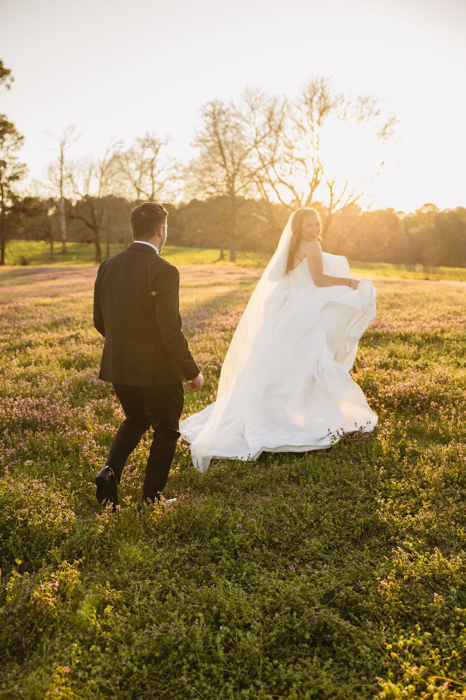 Bride and groom walking through a sunlit meadow at golden hour, wedding veil flowing in the breeze.